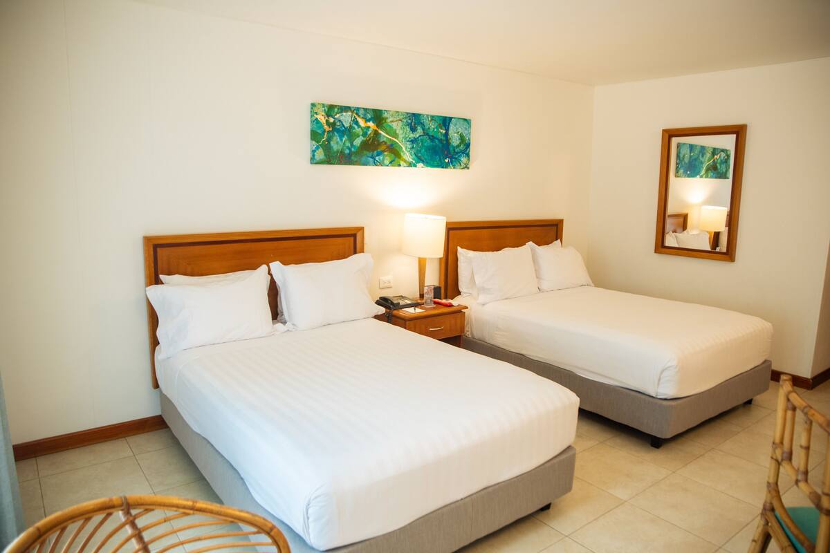 A hotel room with two single beds, a wooden headboard, a nightstand with a lamp, a mirror, and a painting on the wall.