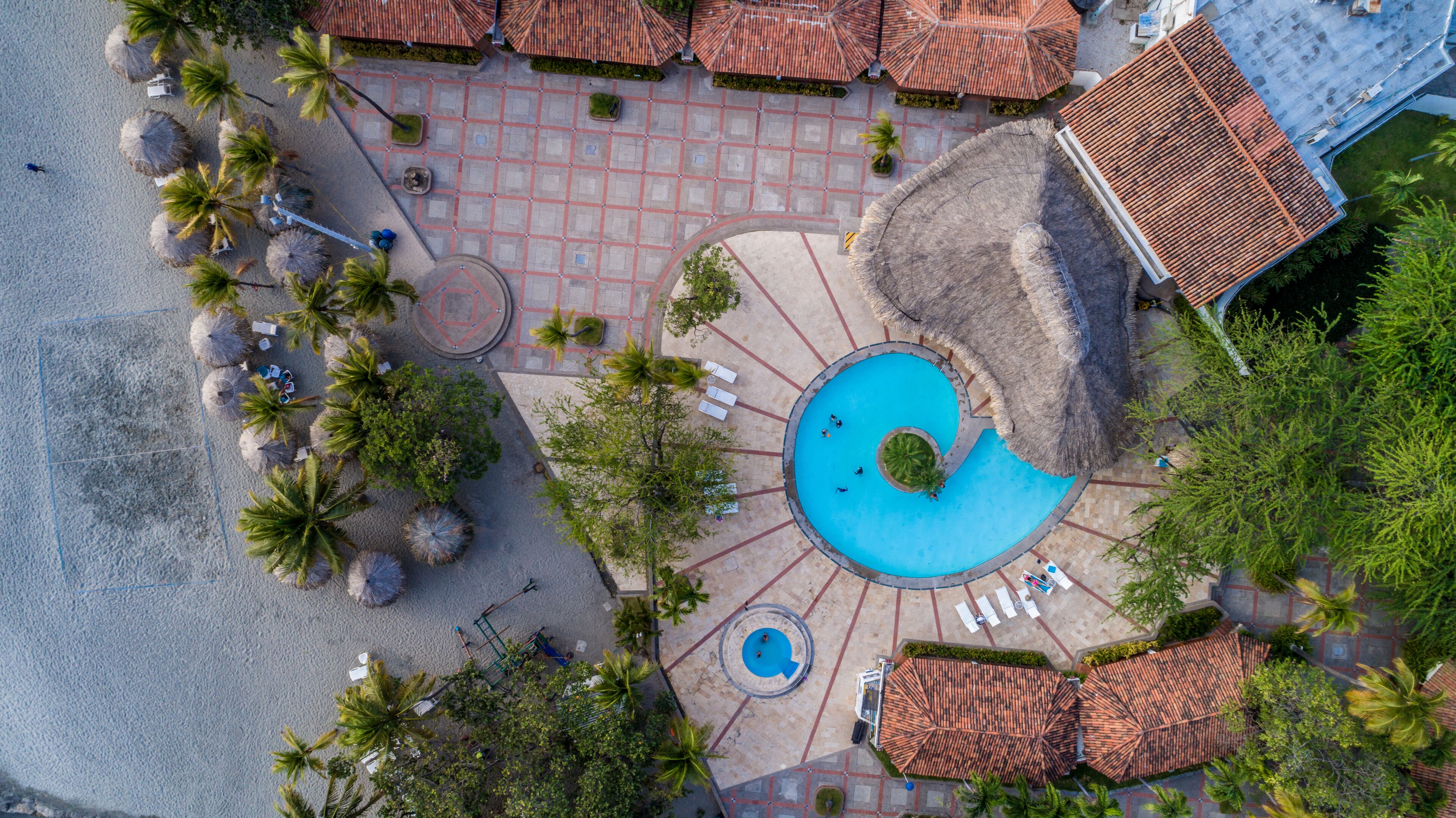 Luchtbeeld van een resort met een centraal gelegen zwembad, palmbomen en betegelde paden.