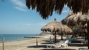 Una playa con sombrillas de paja, sillas reclinables blancas y una vista despejada del mar.