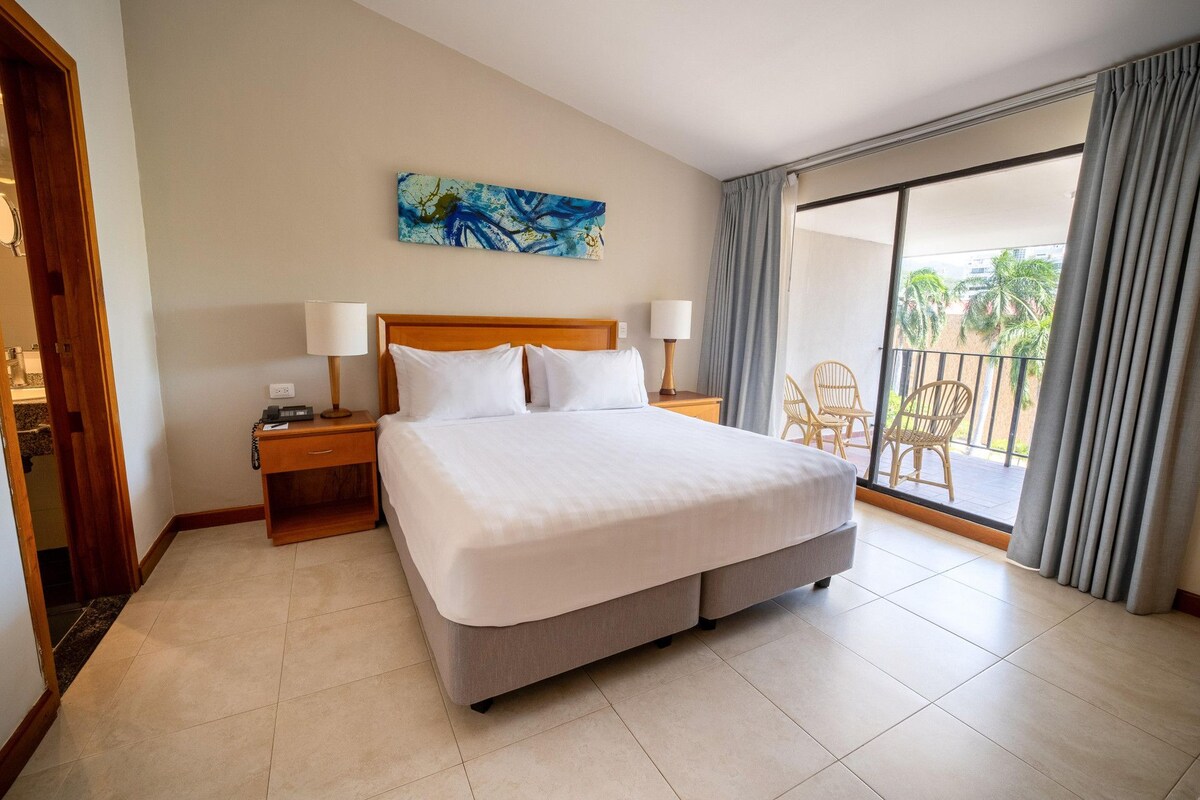 Habitación de hotel con una cama grande, dos mesitas de noche, un cuadro en la pared y un balcón con sillas.