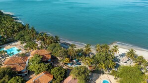 Vista aérea de un complejo turístico frente al mar, con piscina, playa y palmeras.