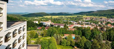 A high-rise building with multiple windows overlooking a town with green hills, a playground, and a swimming pool.