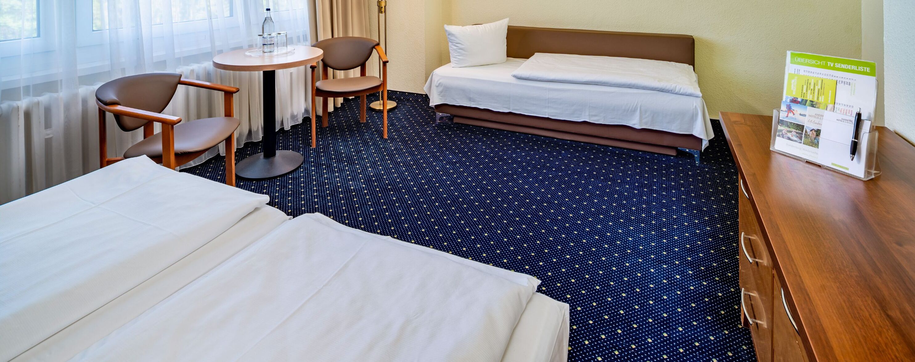 Habitación de hotel con dos camas, una mesita con sillas, un escritorio de madera con portalápices y una ventana con cortinas.
