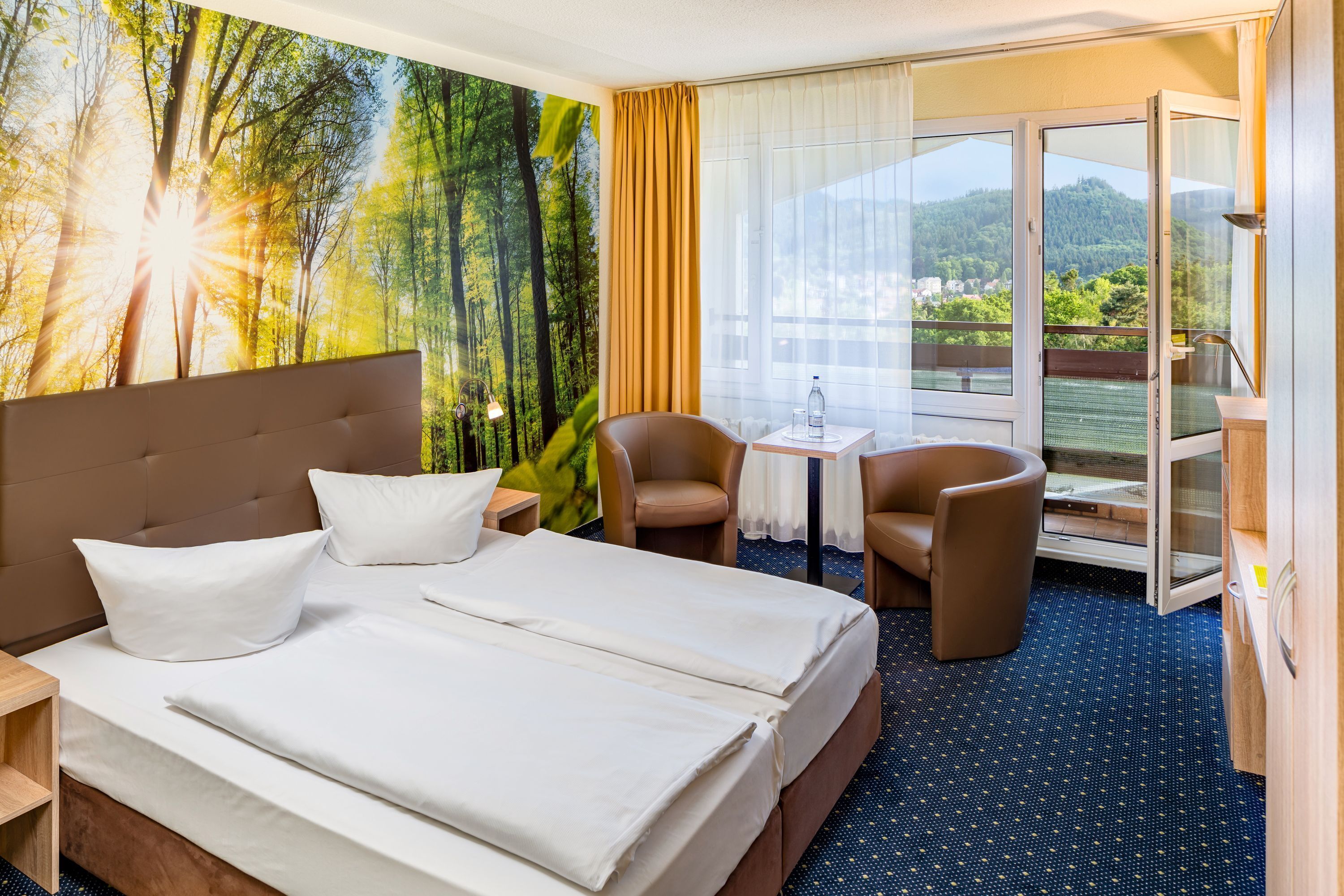 Habitación de hotel con una cama grande, dos sillas, una mesita y vistas a un bosque desde la ventana.