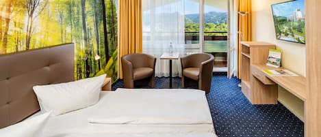 A hotel room with a bed, two chairs, a small table, a desk, and a view of a forest through a window.