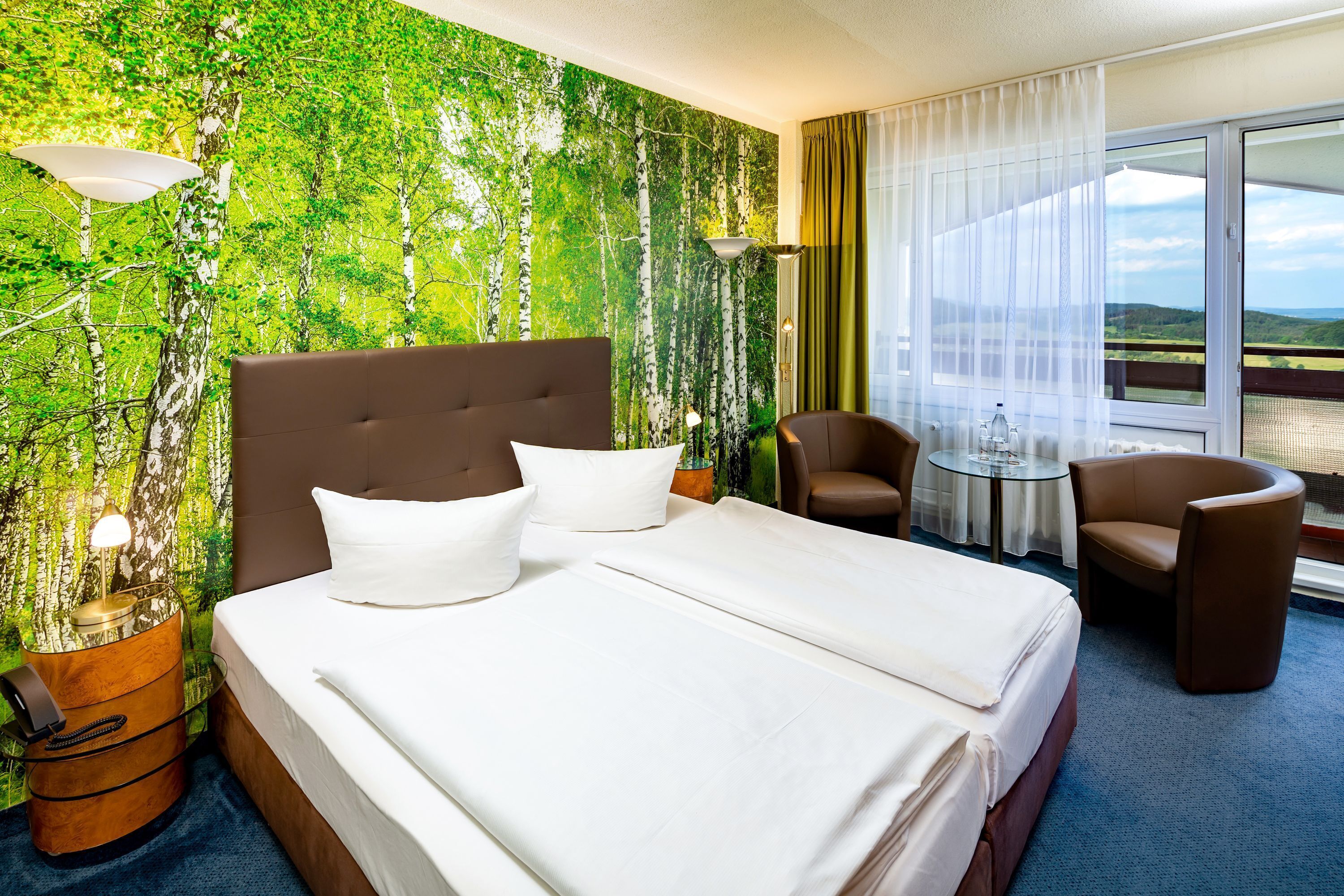 Ett hotellrum med en stor säng, två stolar, ett litet bord och utsikt över omgivningarna.