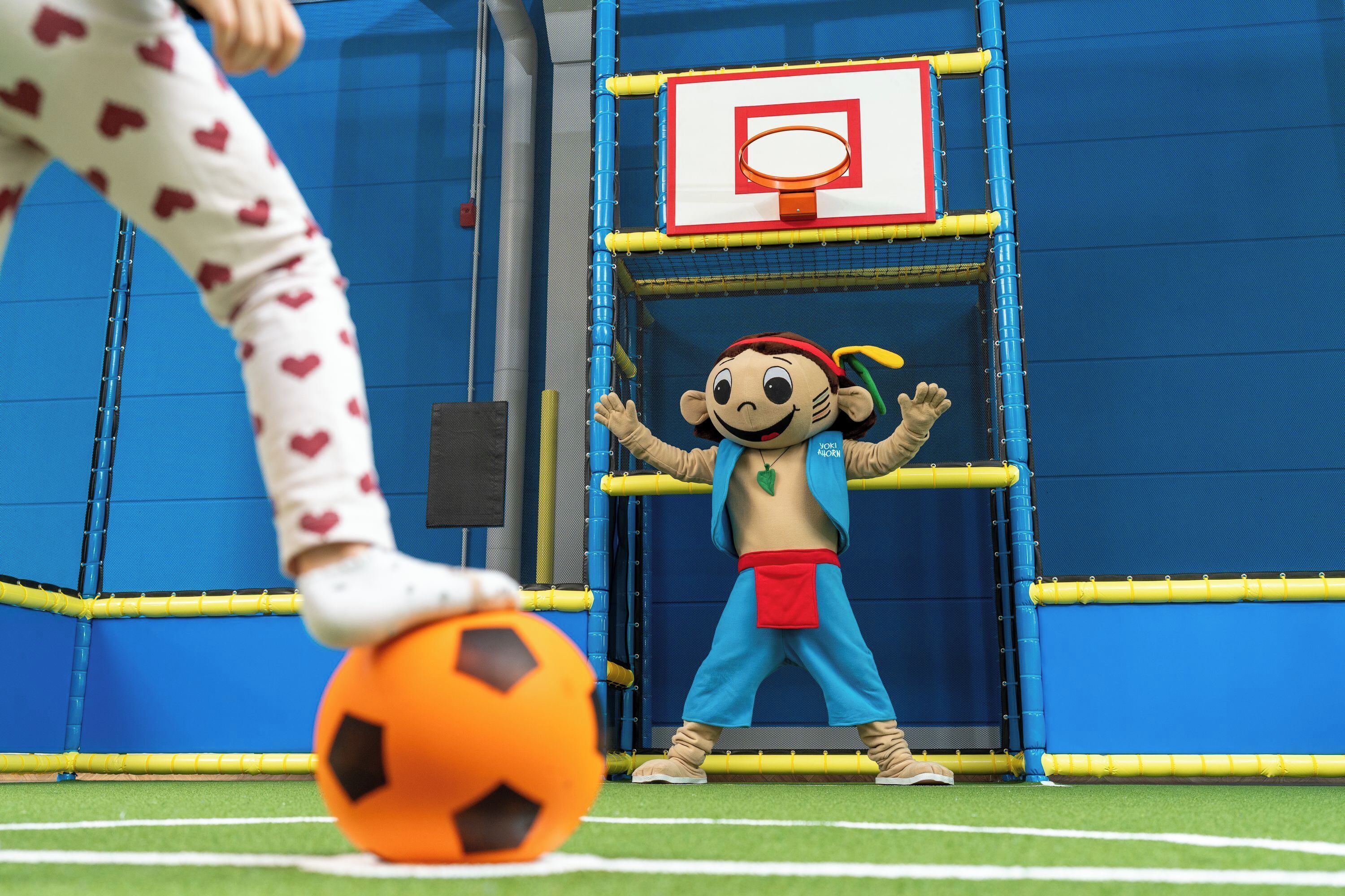 Un personaje promocional con vestimenta azul y roja posado frente a un aro de baloncesto.