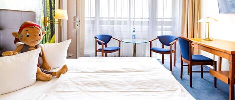A hotel room with a bed, two blue chairs, a small round table with a water bottle, and a desk with a lamp.
