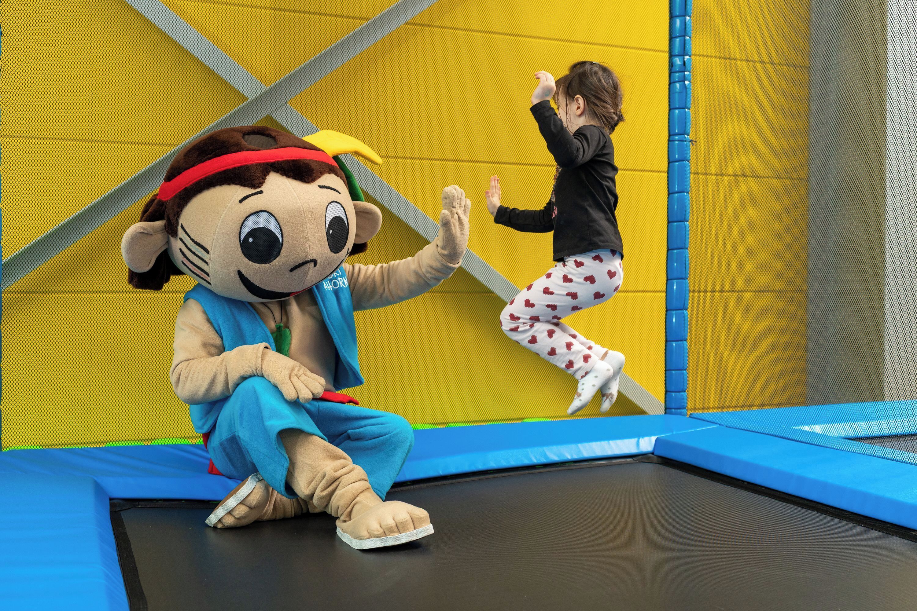 A mascot sitting on a trampoline with a child jumping nearby.
