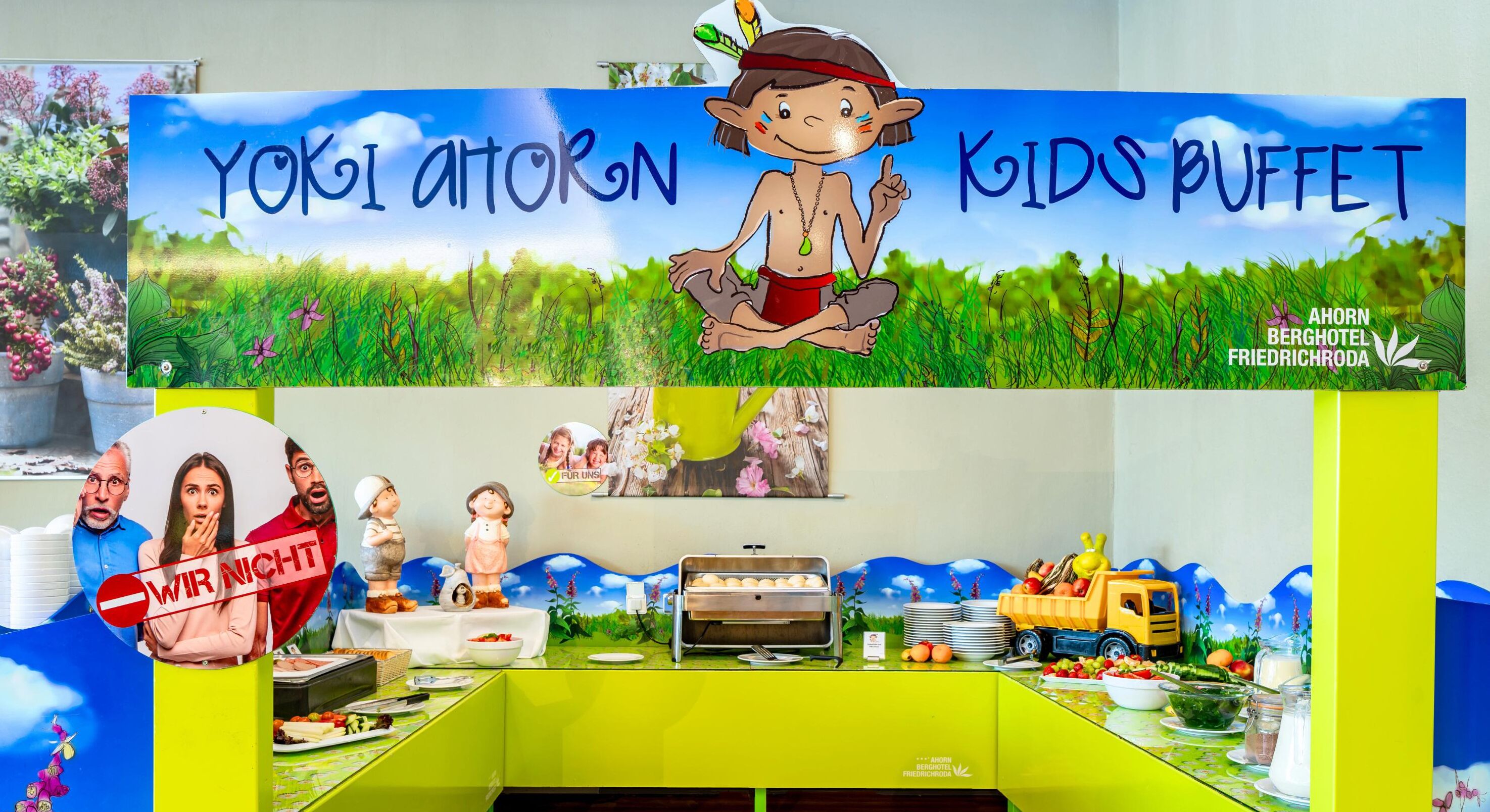 A children's buffet area with a sign reading "YOKI AHTORON KIDS BUFFET" and a cartoon character. There are food items, a yellow truck decoration, and a table with plates and utensils.
