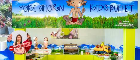 Zona de bufé infantil con un letrero que dice "YOKI AHTORON KIDS BUFFET" y un personaje de caricatura. Se observan alimentos, una decoración de camión amarillo y una mesa con platos y cubiertos.