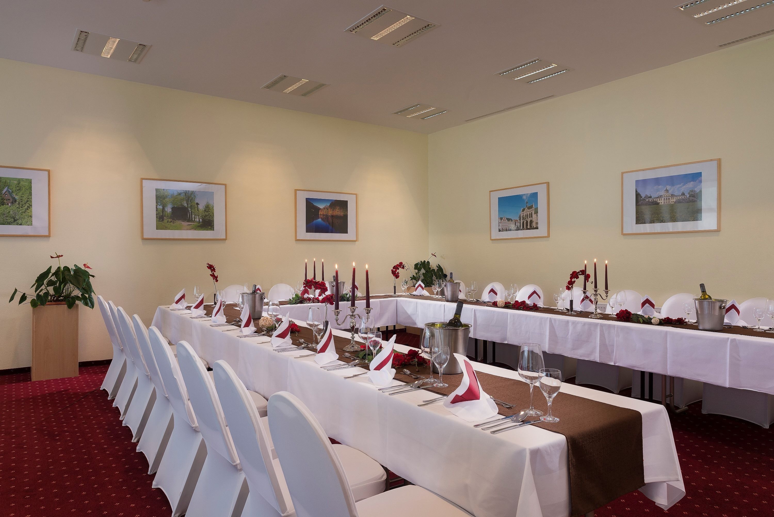 A conference room with a long table set for a formal event, featuring white chairs, a white tablecloth, and red floral decorations.