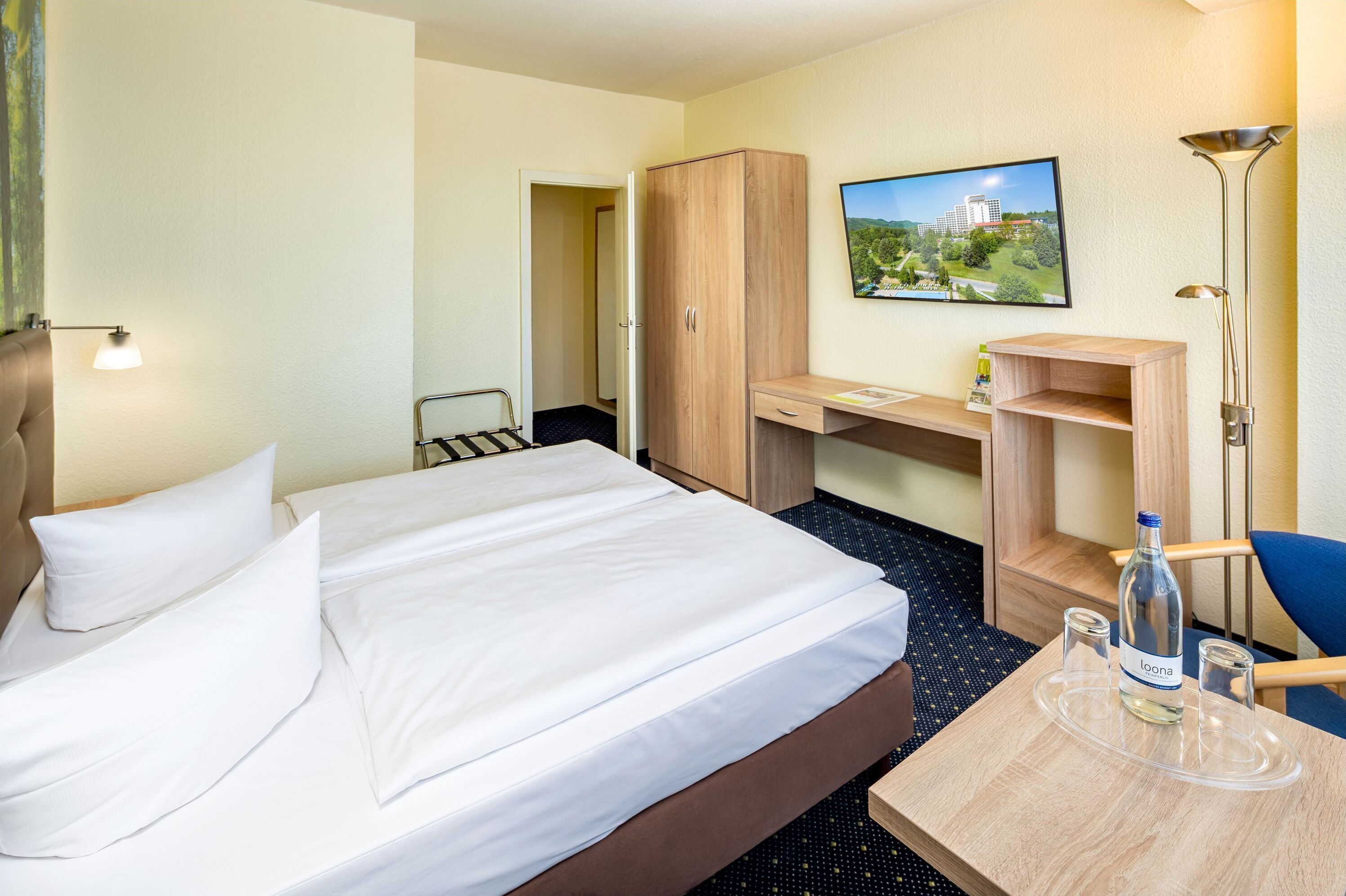 A hotel room with a bed, a desk, a wardrobe, a chair, and a table with a bottle and glasses.