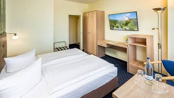 Quarto de hotel com uma cama, uma escrivaninha, um guarda-roupa, uma cadeira e uma mesa com garrafa e copos.