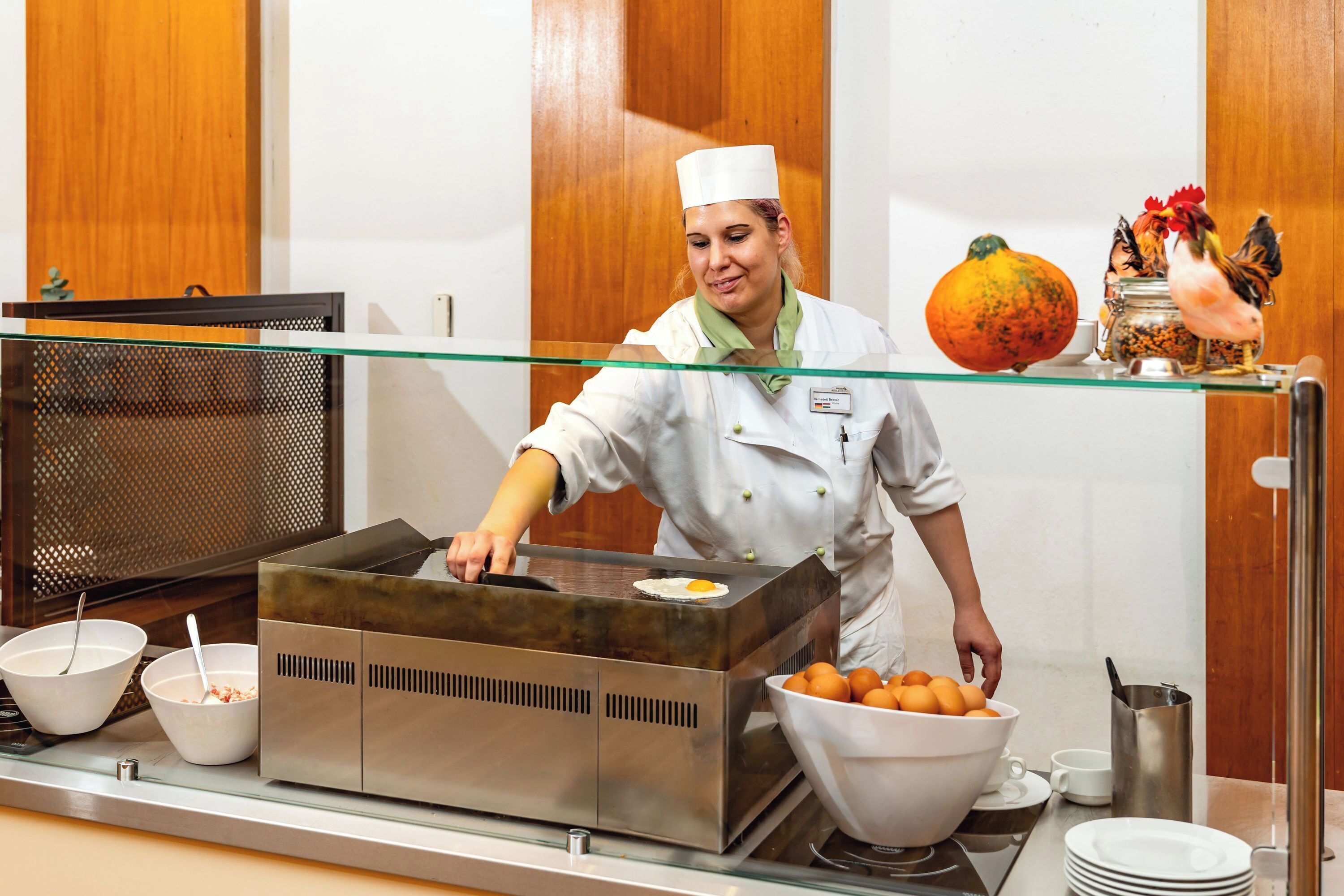 Um chef numa cozinha com estação de cozinha em aço inoxidável, tigelas com ingredientes e um galo decorativo numa prateleira.