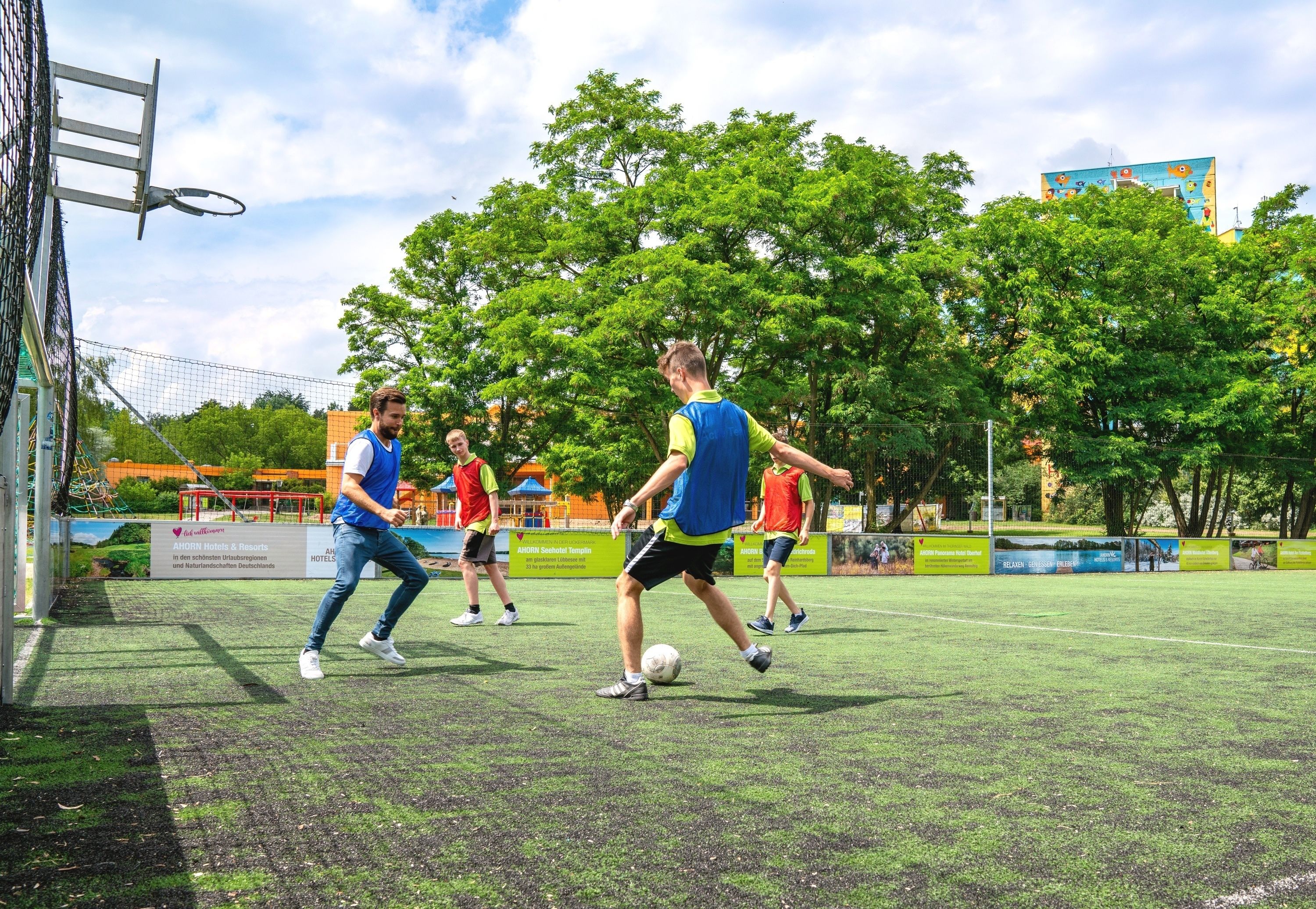 Un grupo de personas jugando al fútbol en una cancha al aire libre con un aro de baloncesto y árboles de fondo.