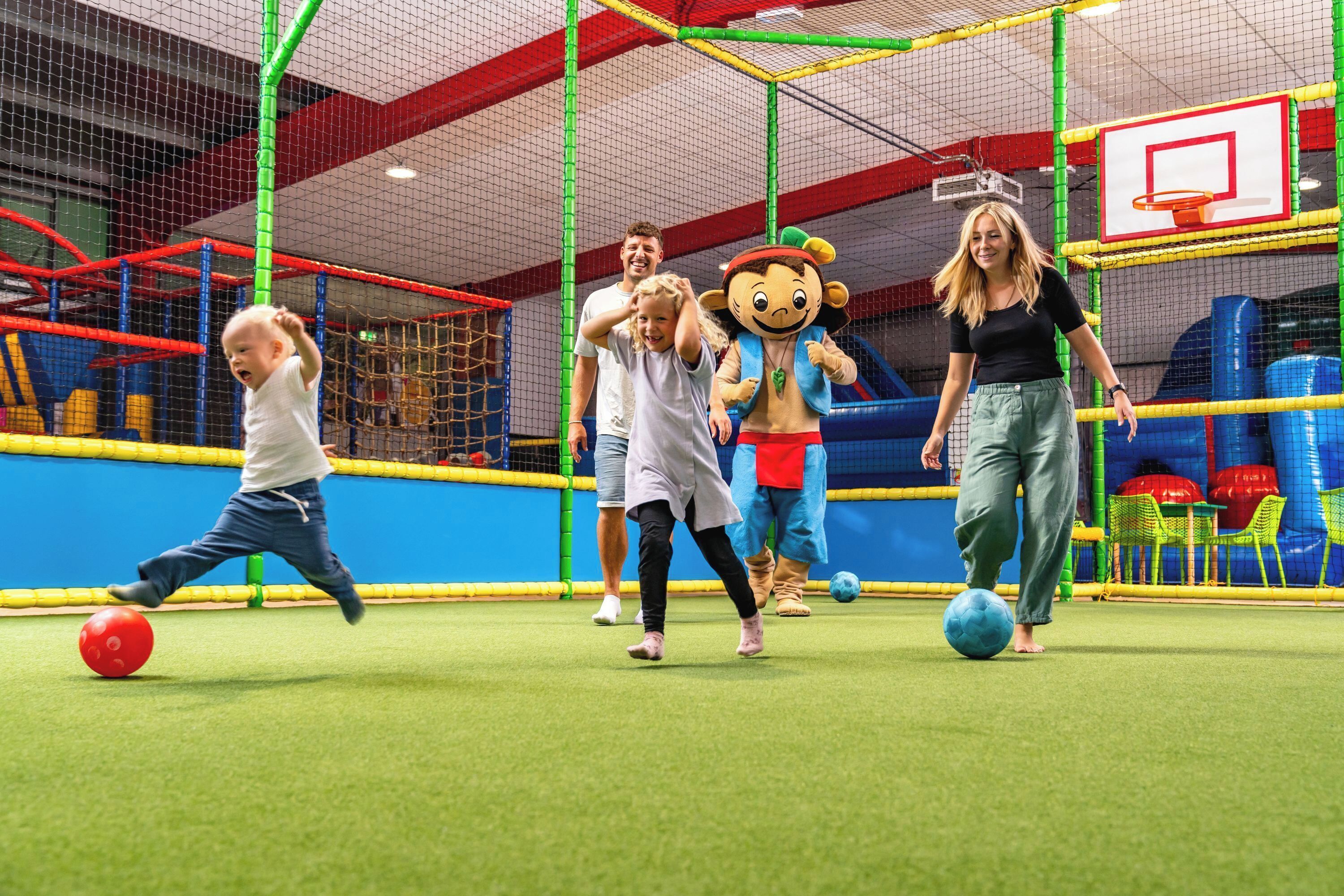 Zona de juegos con un aro de baloncesto, estructuras de juego con redes y un personaje mascota.