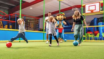Zona de juegos con un aro de baloncesto, estructuras de juego con redes y un personaje mascota.