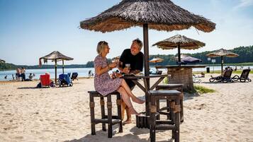 Un café situé en bord de mer, avec des tables en bois et des parasols en chaume, offrant une vue sur une plage de sable où l’on aperçoit des personnes et des bateaux au loin.