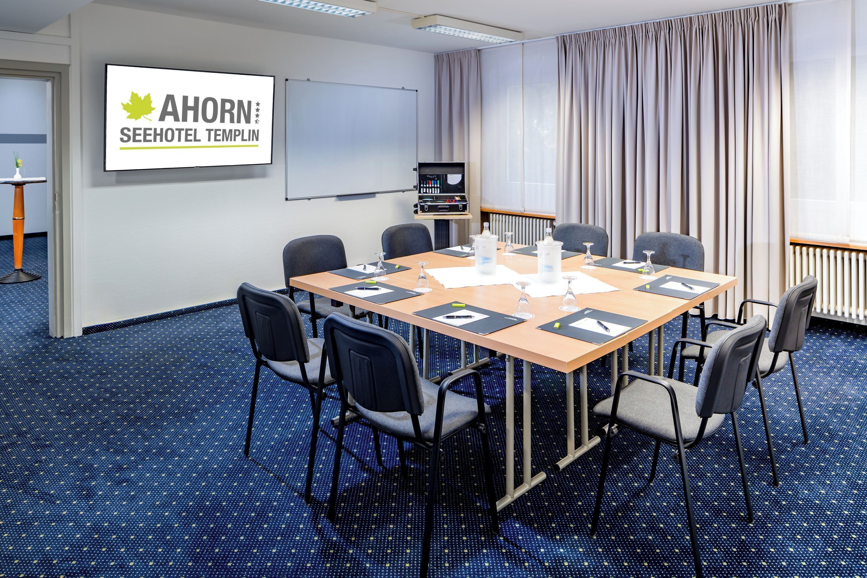 Un salón de conferencias con una larga mesa, seis sillas, una pantalla de proyector y un cartel que dice "AHORN SEEHOTEL TEMPLIN".