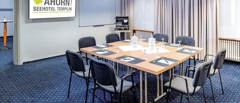 Un salón de conferencias con una larga mesa, seis sillas, una pantalla de proyector y un cartel que dice "AHORN SEEHOTEL TEMPLIN".