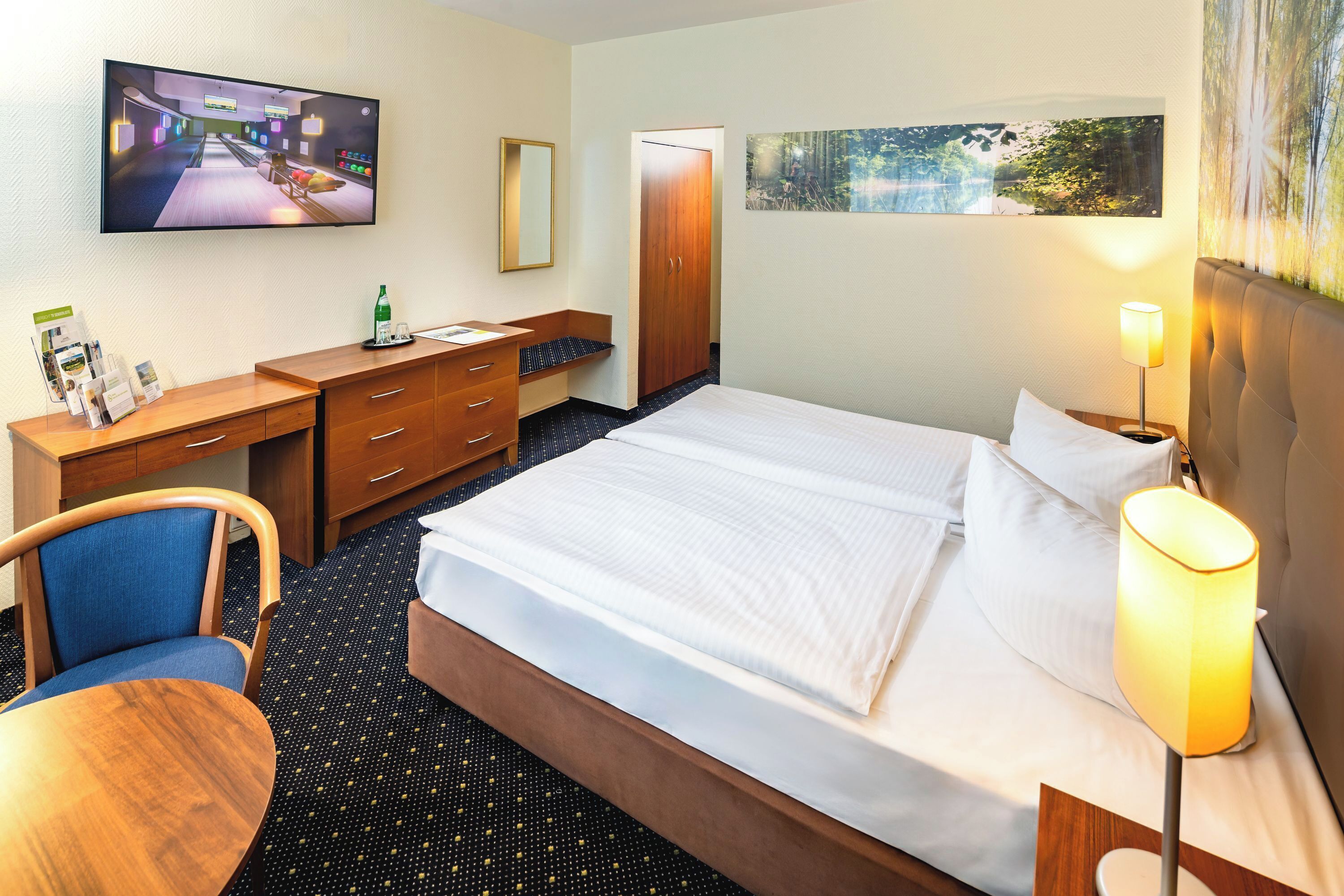 A hotel room with a bed, a desk with a chair, a television, and a painting on the wall.