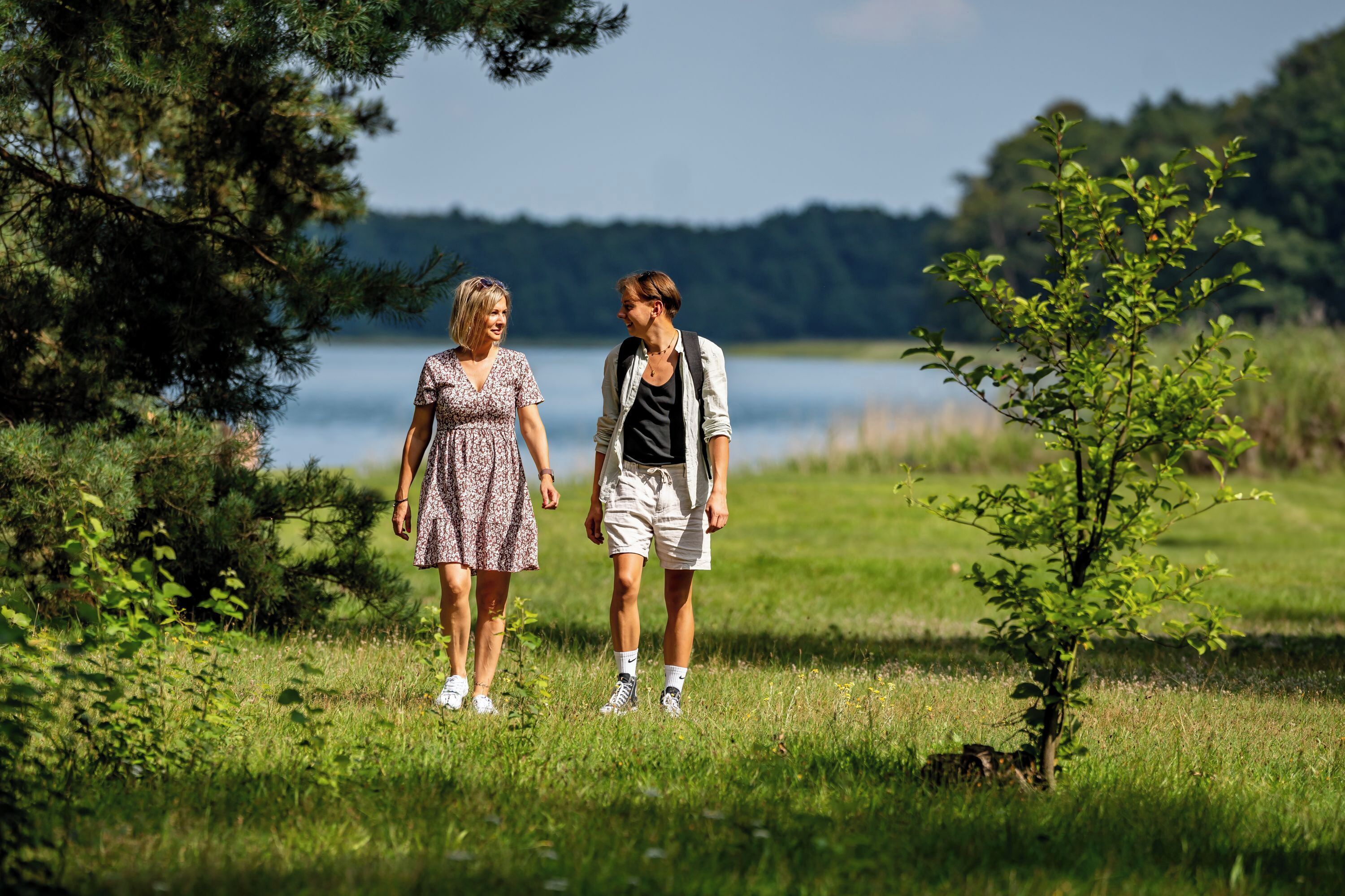 Two people walking in a grassy area with trees and a body of water in the background.