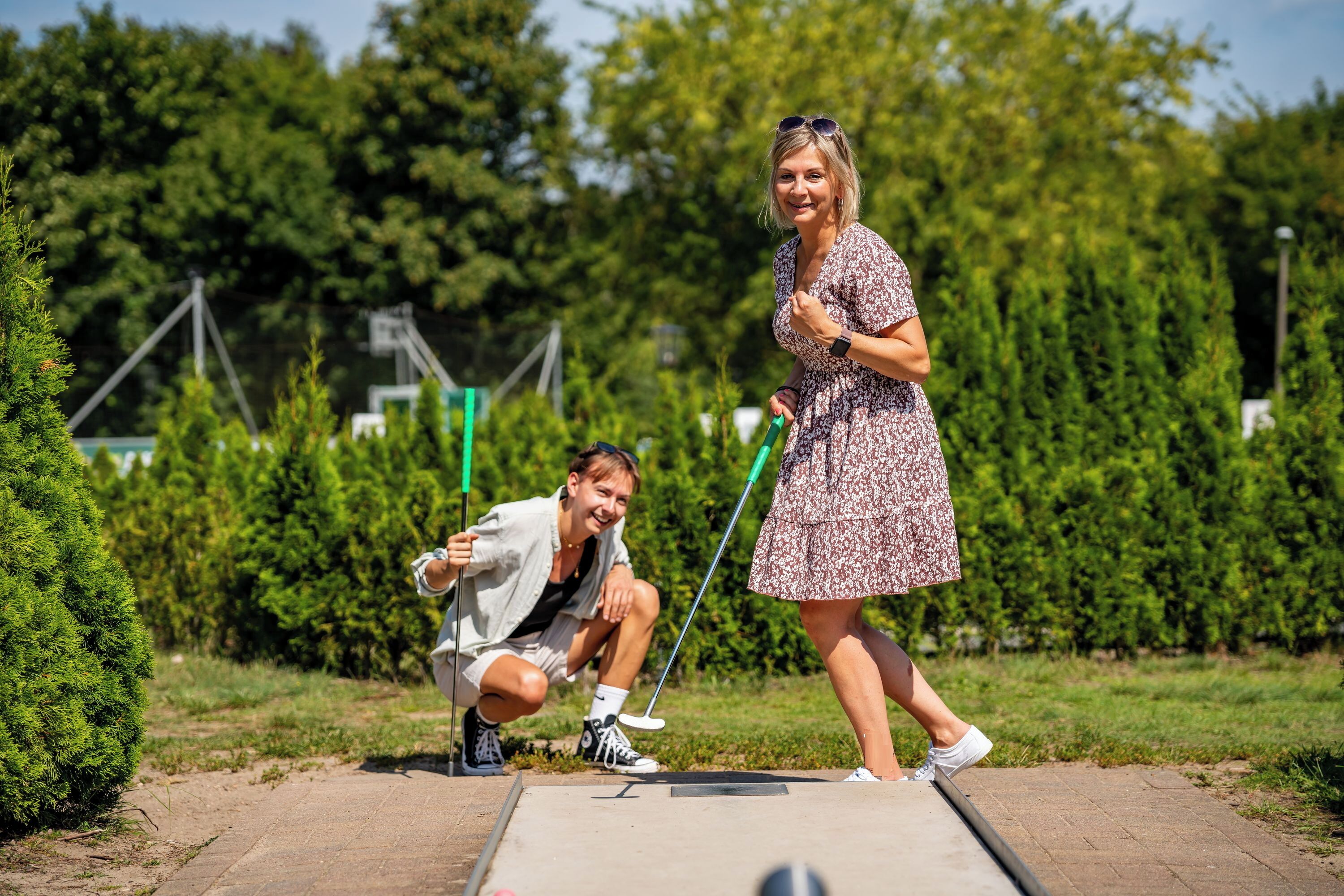 A person in a patterned dress and another in casual attire at a golf driving range with trees and greenery in the background.