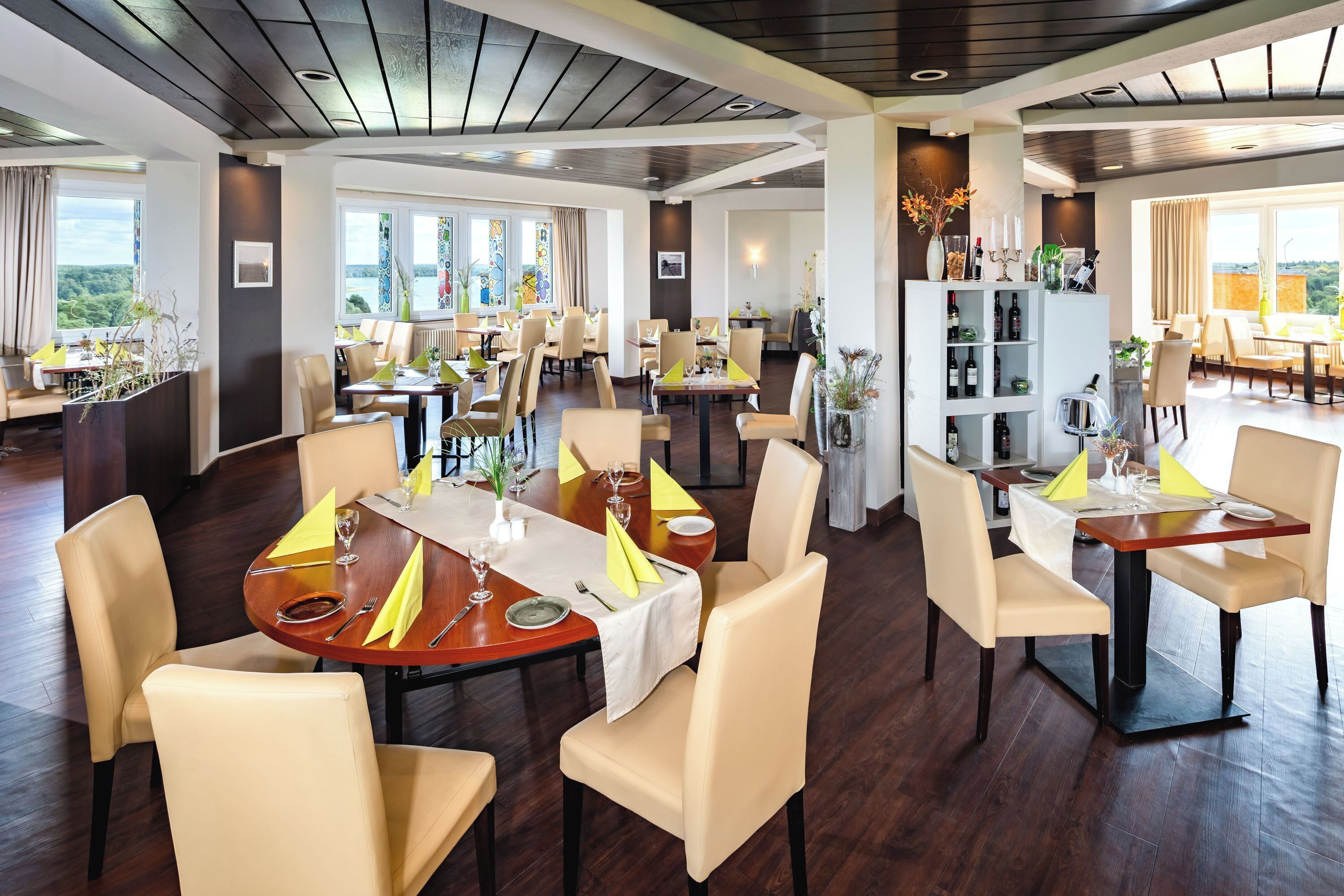 Un comedor amplio con varias mesas redondas, cada una con servilletas amarillas, copas de vino y cubiertos. La habitación tiene pisos de madera oscura, ventanas grandes con cortinas y vistas a follaje exterior.
