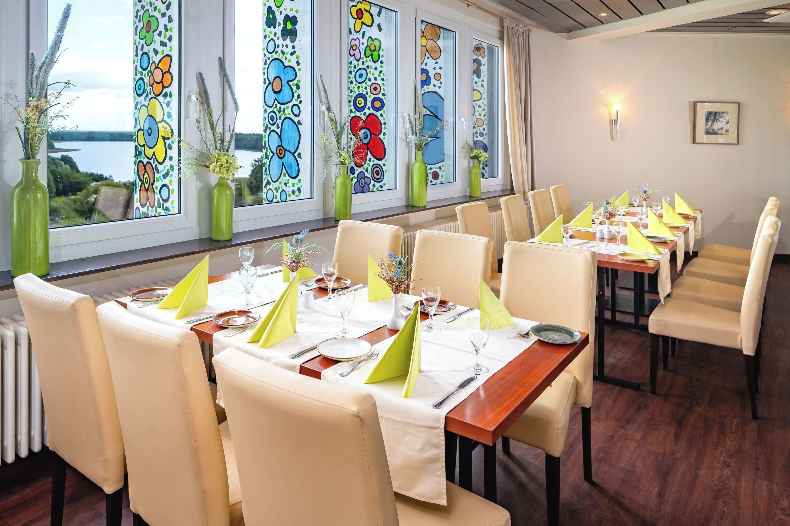 A restaurant with a long dining table set for a meal, large windows with colorful stained glass, and a view of the outdoors.