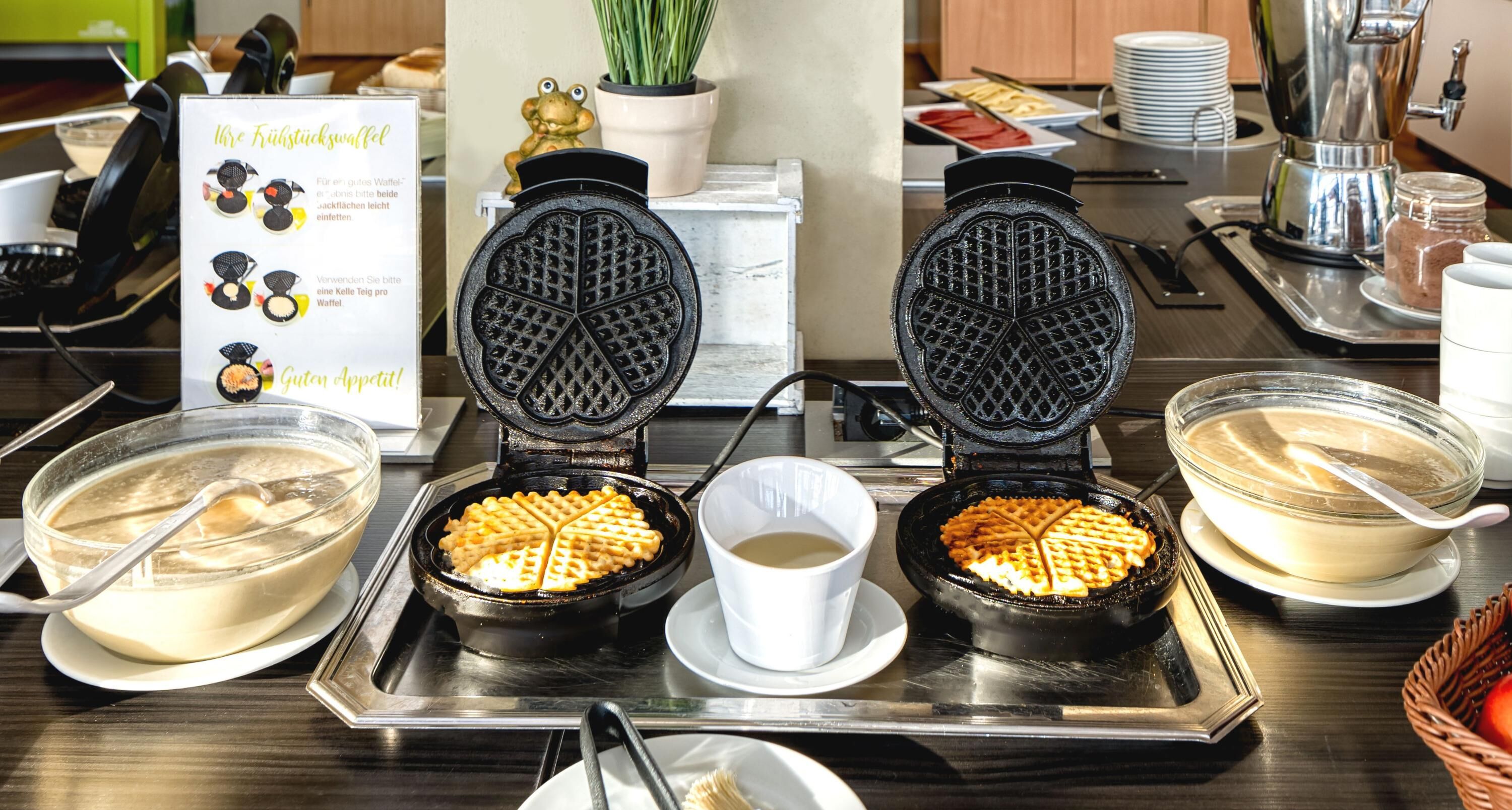 A waffle station with two waffle irons, a bowl of batter, a cup of coffee, and a sign with instructions.
