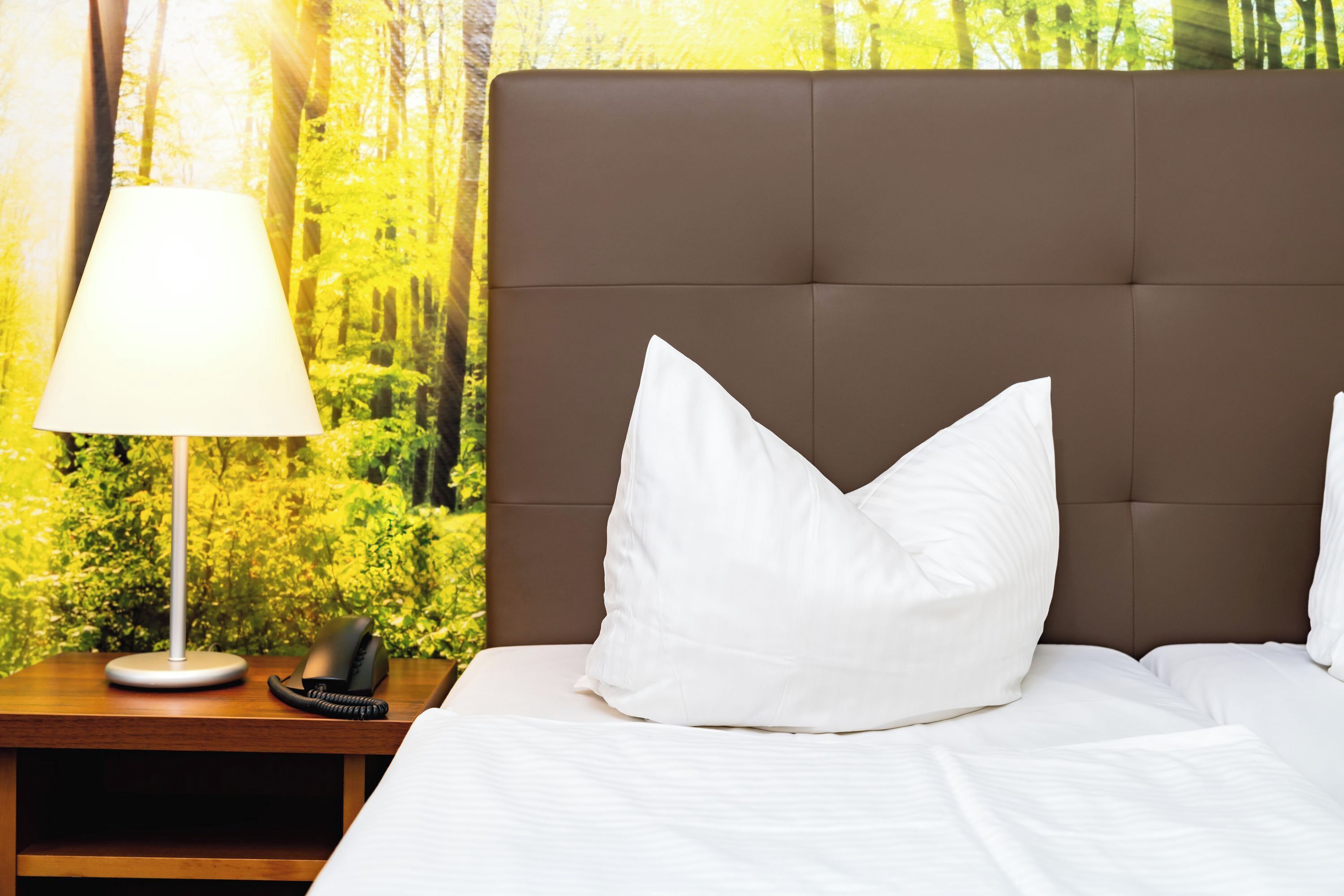 Una cama bien hecha con ropa de cama y almohadas blancas, una lámpara de noche y un teléfono sobre una mesita de madera.