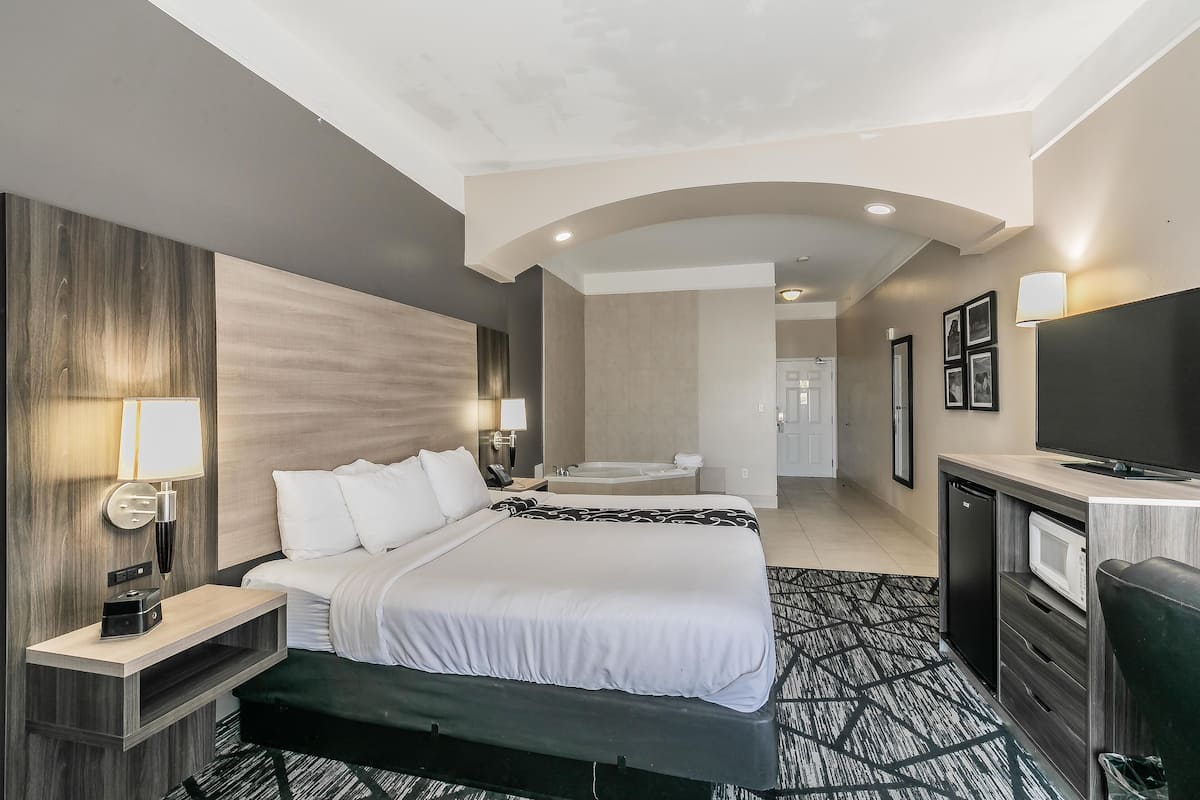 Een hotelkamer met een groot bed, nachtkastje, lamp, televisie en uitzicht op de gang.