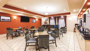 A dining area with multiple tables and chairs, a television mounted on the wall, and a kitchen area with appliances.
