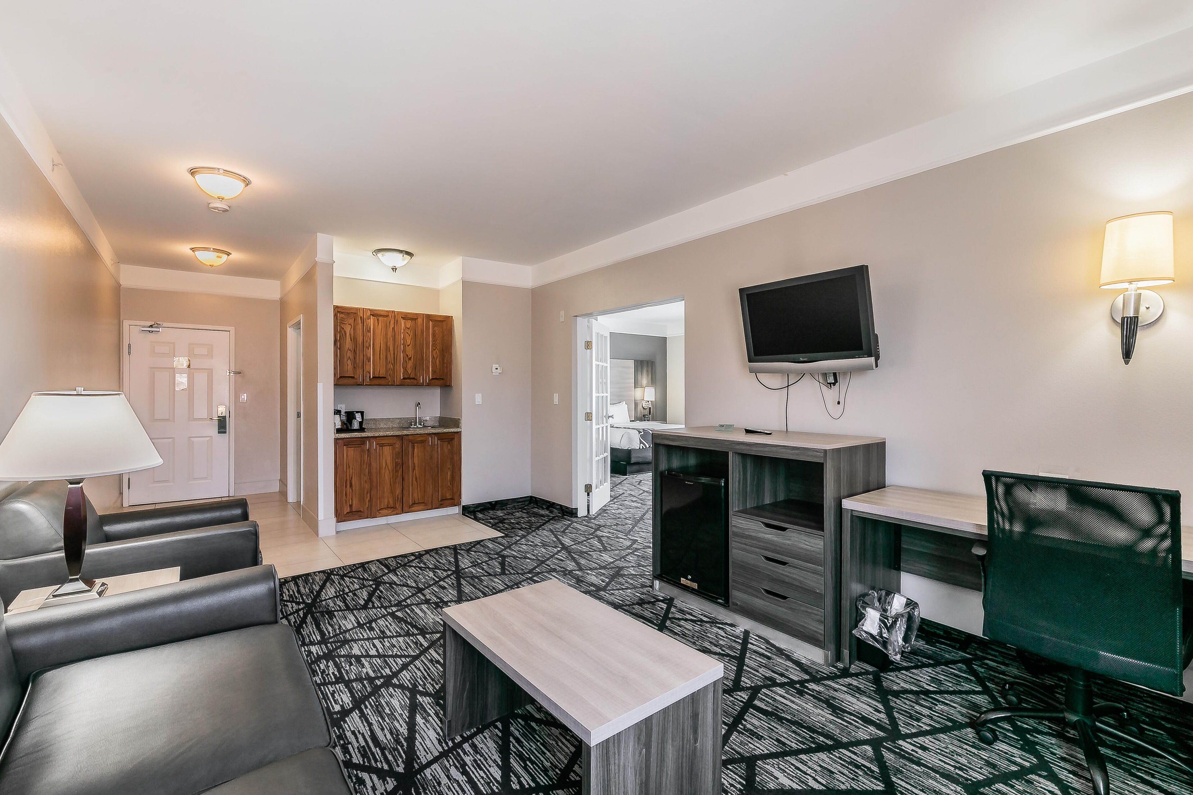A modern hotel room with a sofa, a desk, a TV, and a kitchen area.
