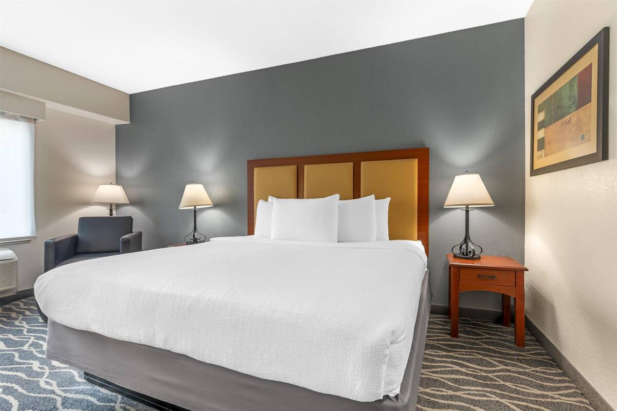 A hotel room with a large bed, two bedside lamps, a small table with a lamp, a chair, and a painting on the wall.