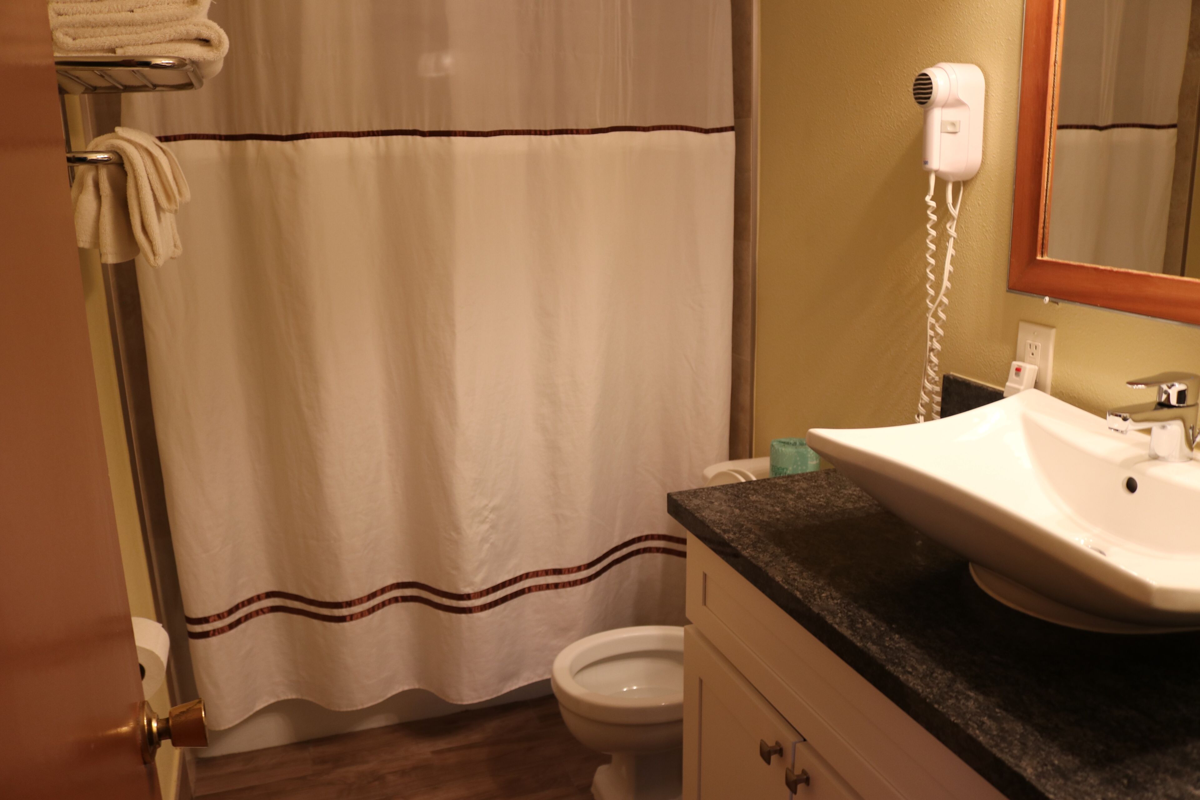 A bathroom with a white shower curtain, a white toilet, a sink with a black countertop, and a hairdryer on the wall.