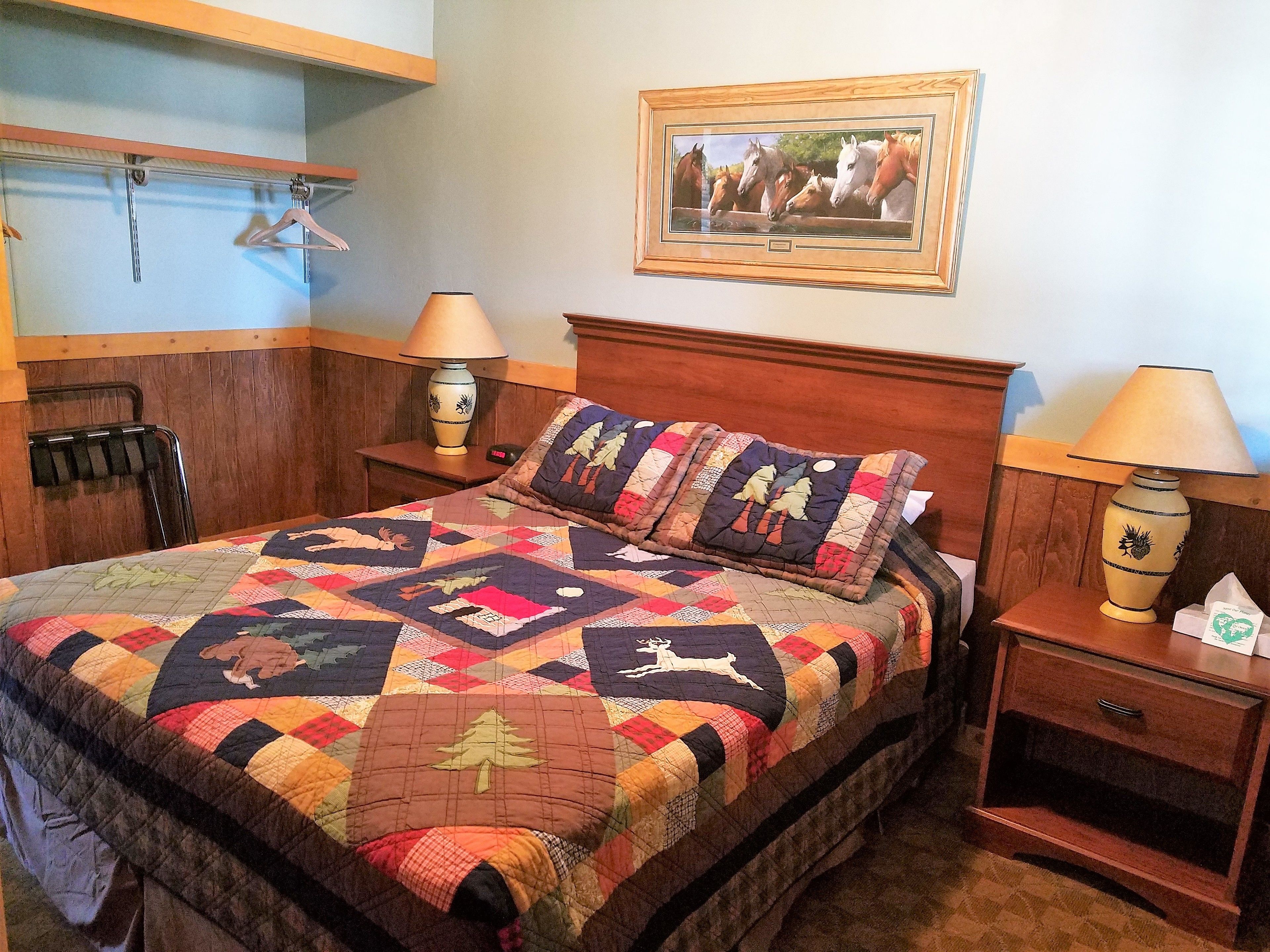 A neatly made bed with a colorful quilt, two bedside tables with lamps, a wooden headboard, a coat rack, and a framed picture on the wall.
