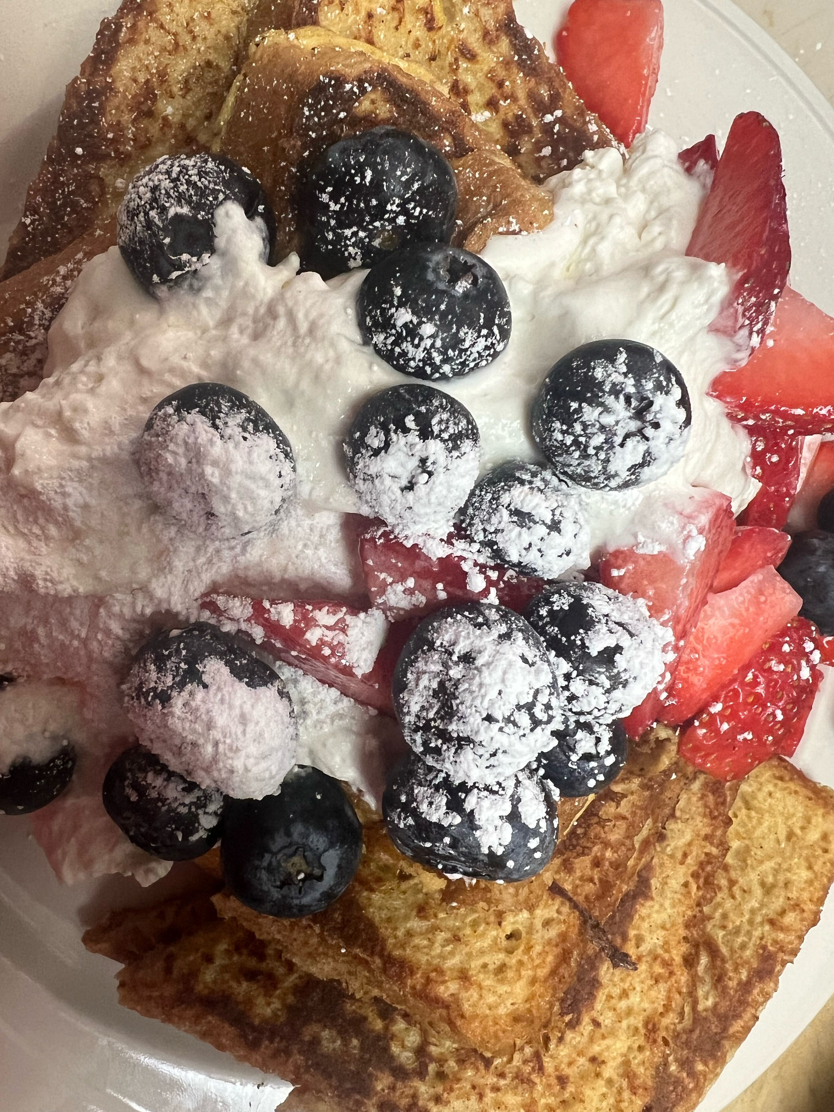 A plate of French toast with blueberries and strawberries, topped with whipped cream and powdered sugar.