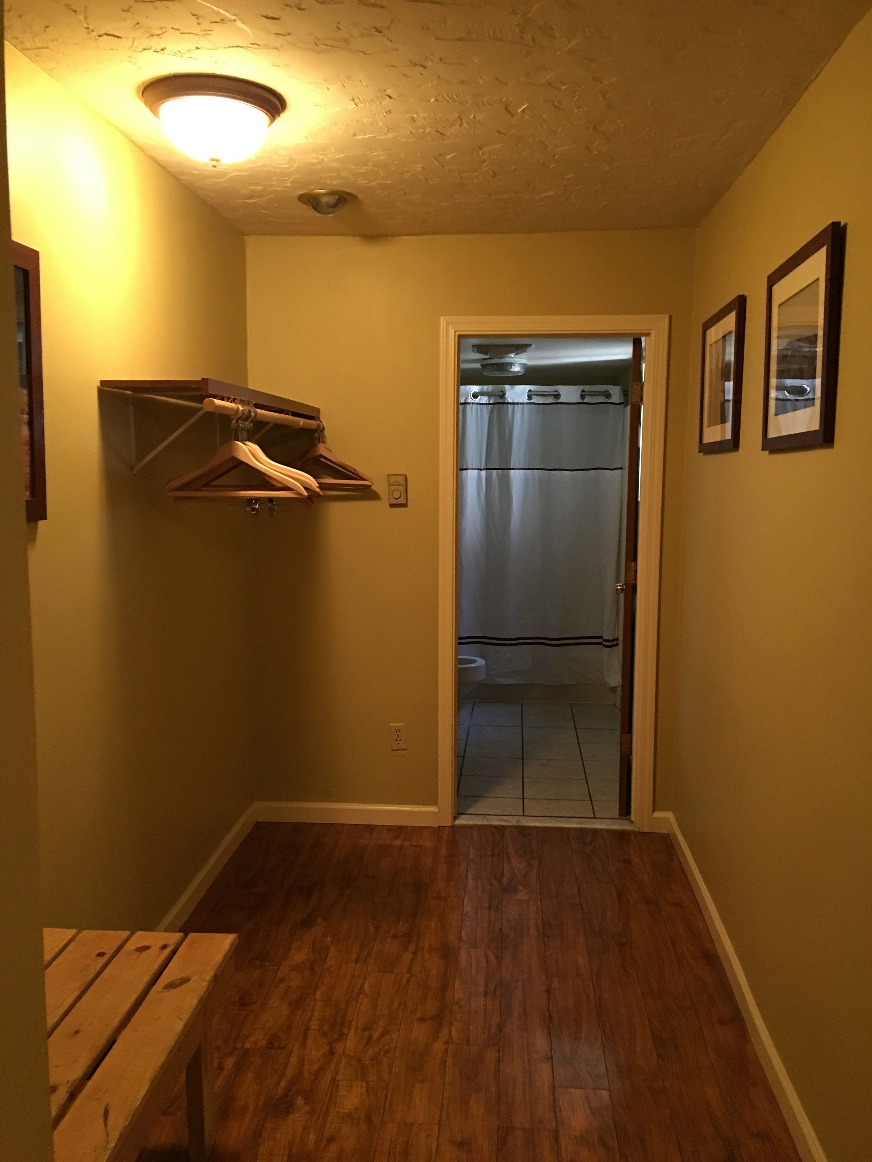 A hallway with wooden flooring, a ceiling light, a coat rack, and a door leading to a bathroom with a shower curtain.