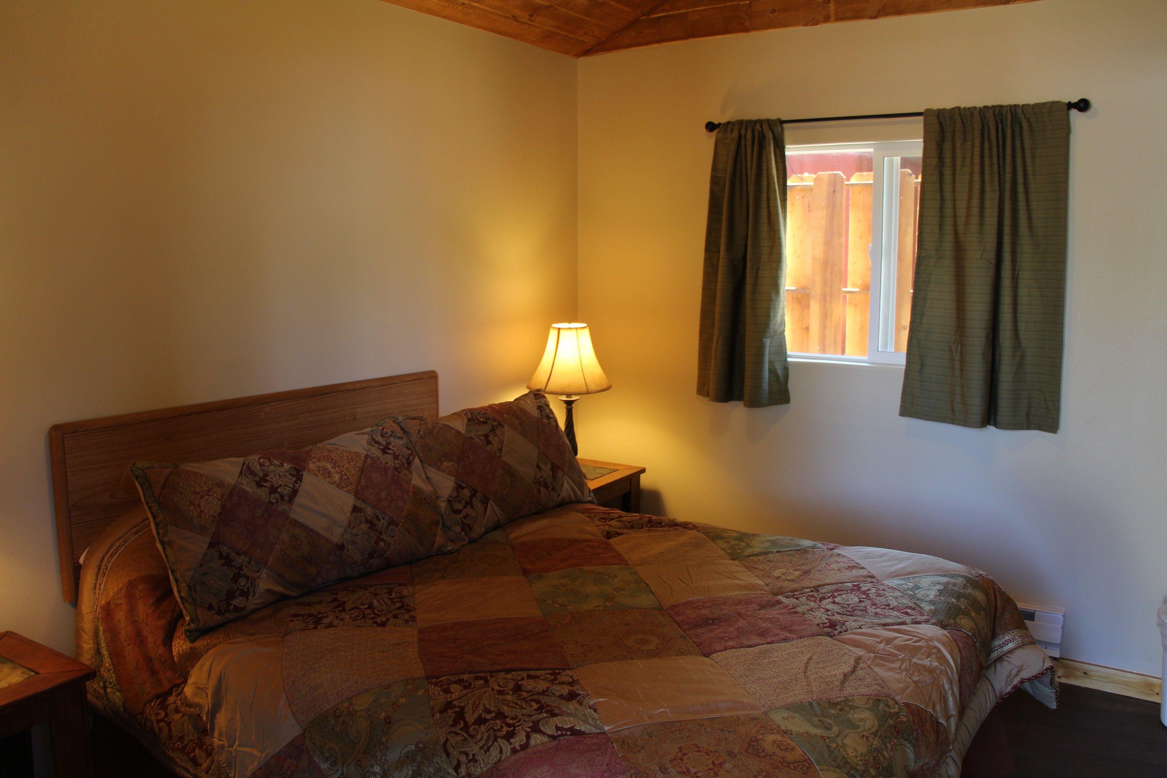 A bedroom with a wooden headboard, a quilted bedspread, a bedside lamp, and a window with curtains.