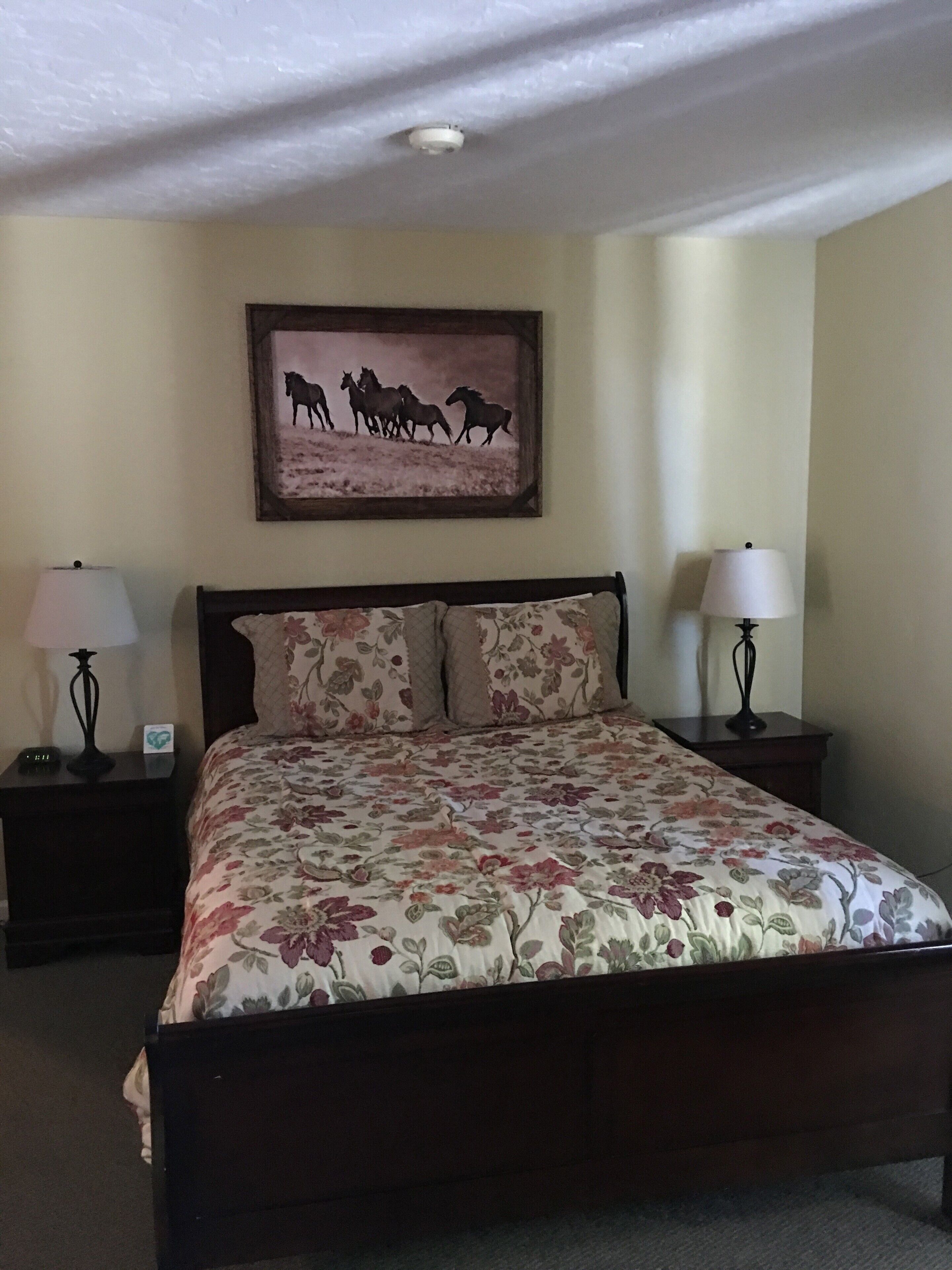Une chambre à coucher avec une couvre-lit à motifs floraux, deux lampes de chevet, un tableau représentant des chevaux accroché au mur et un luminaire au plafond.