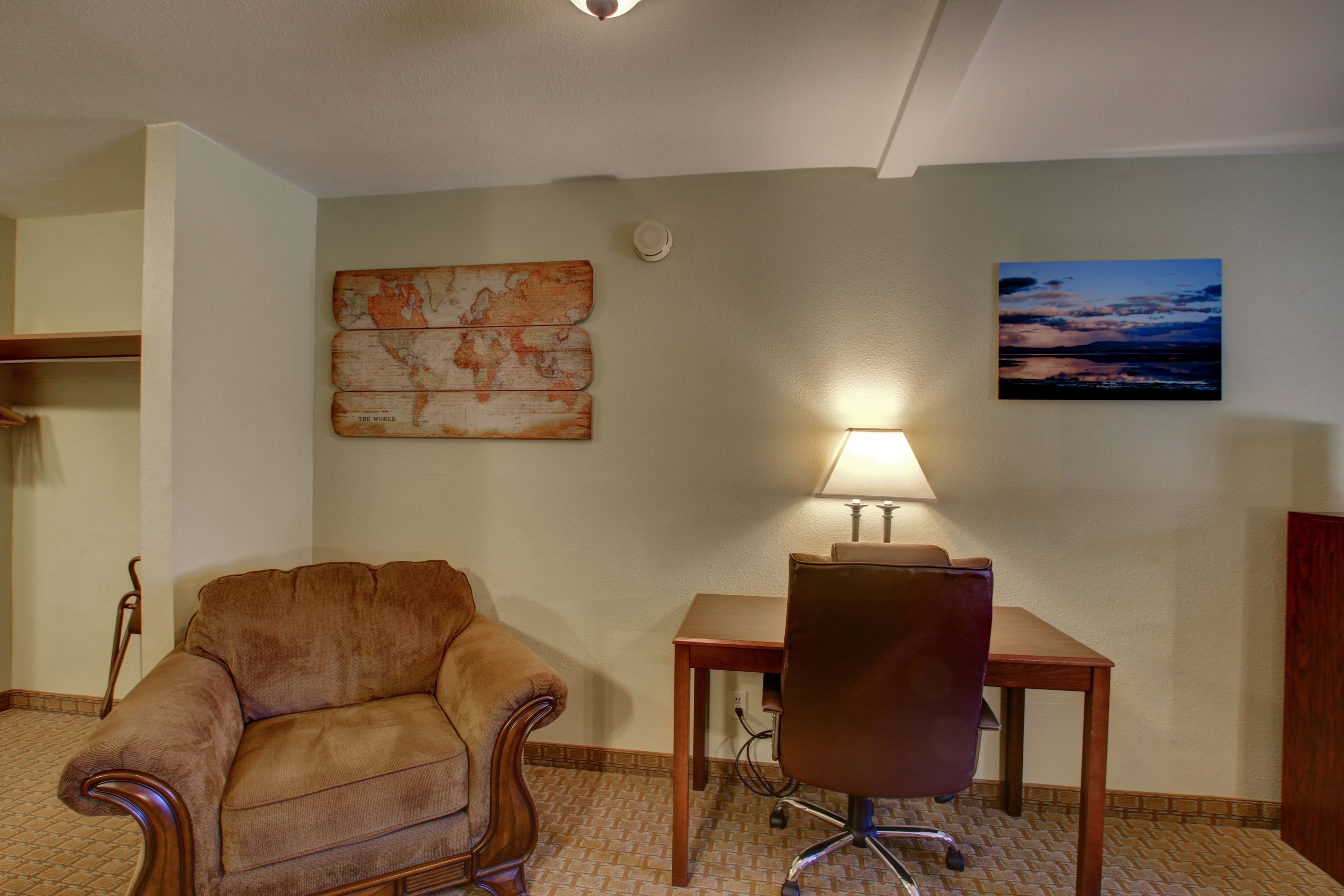 A room with a brown armchair, a wooden table with a lamp, a world map, and a sunset painting on the wall.