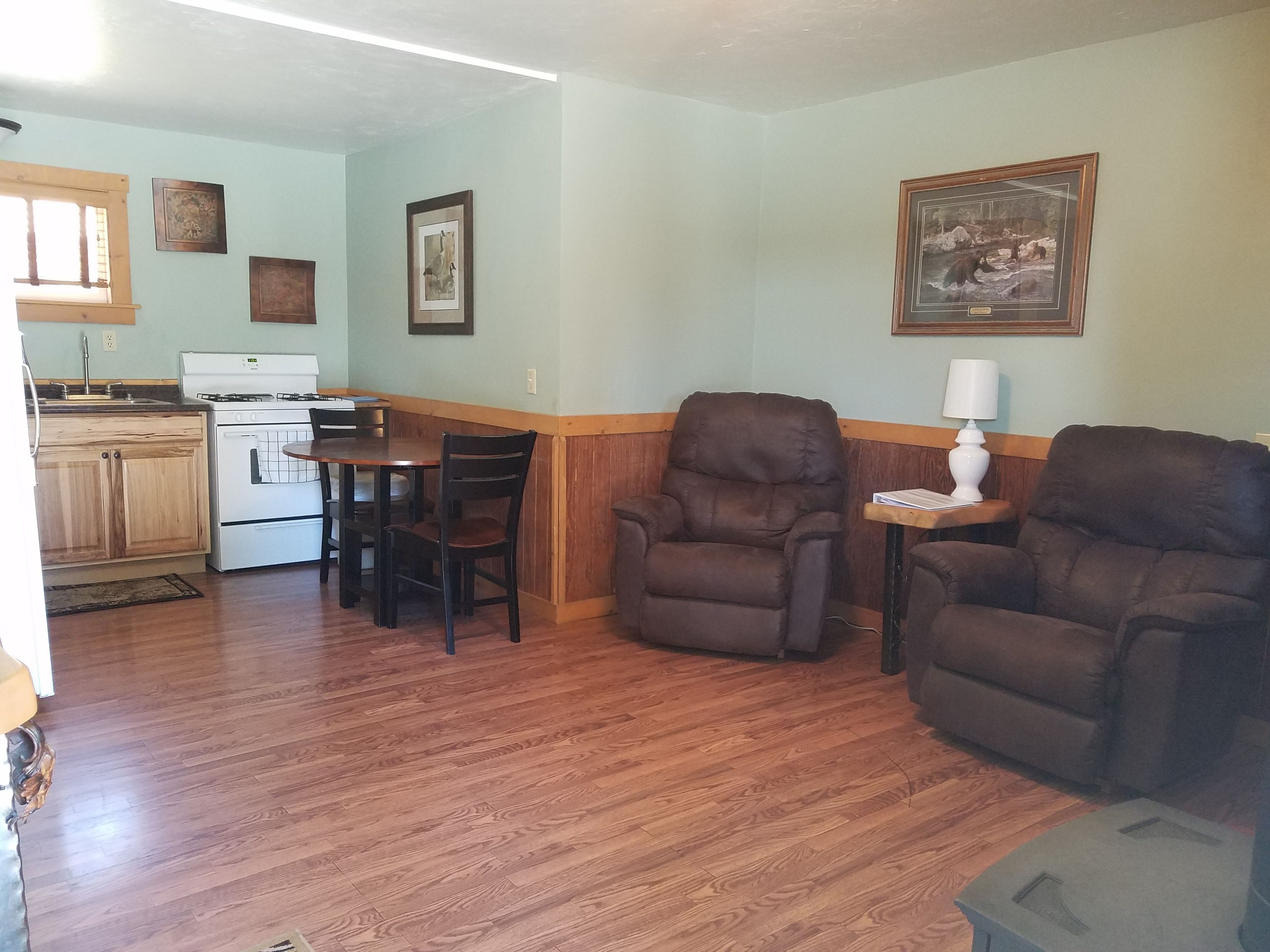 Una cocina con armarios de madera, una estufa y una mesa de comedor con sillas. Hay dos sillones y una lámpara sobre una mesita.