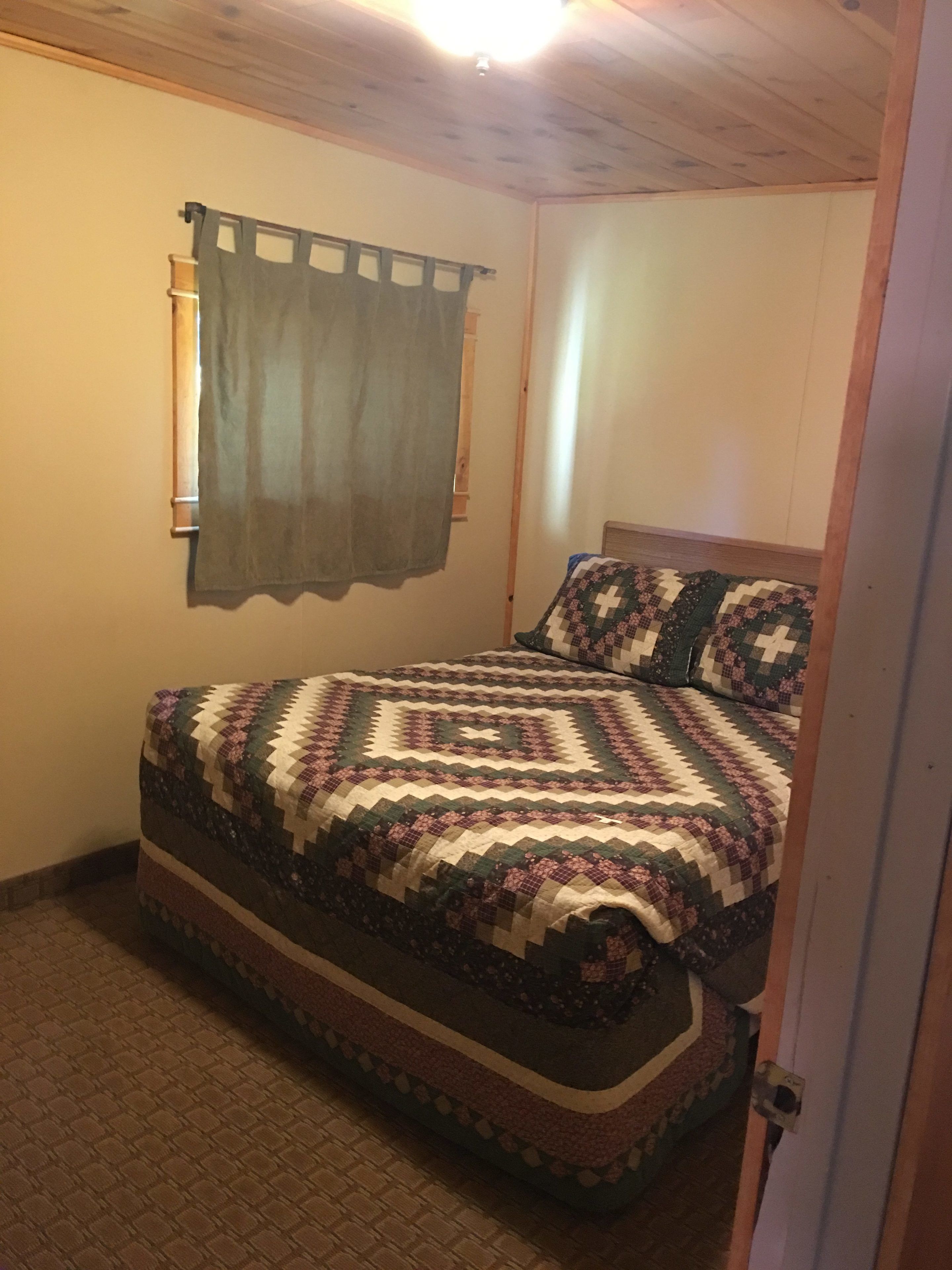 Une chambre avec un plafond en bois, un lit recouvert d’une courtepointe à motifs, une fenêtre avec un rideau et une porte.