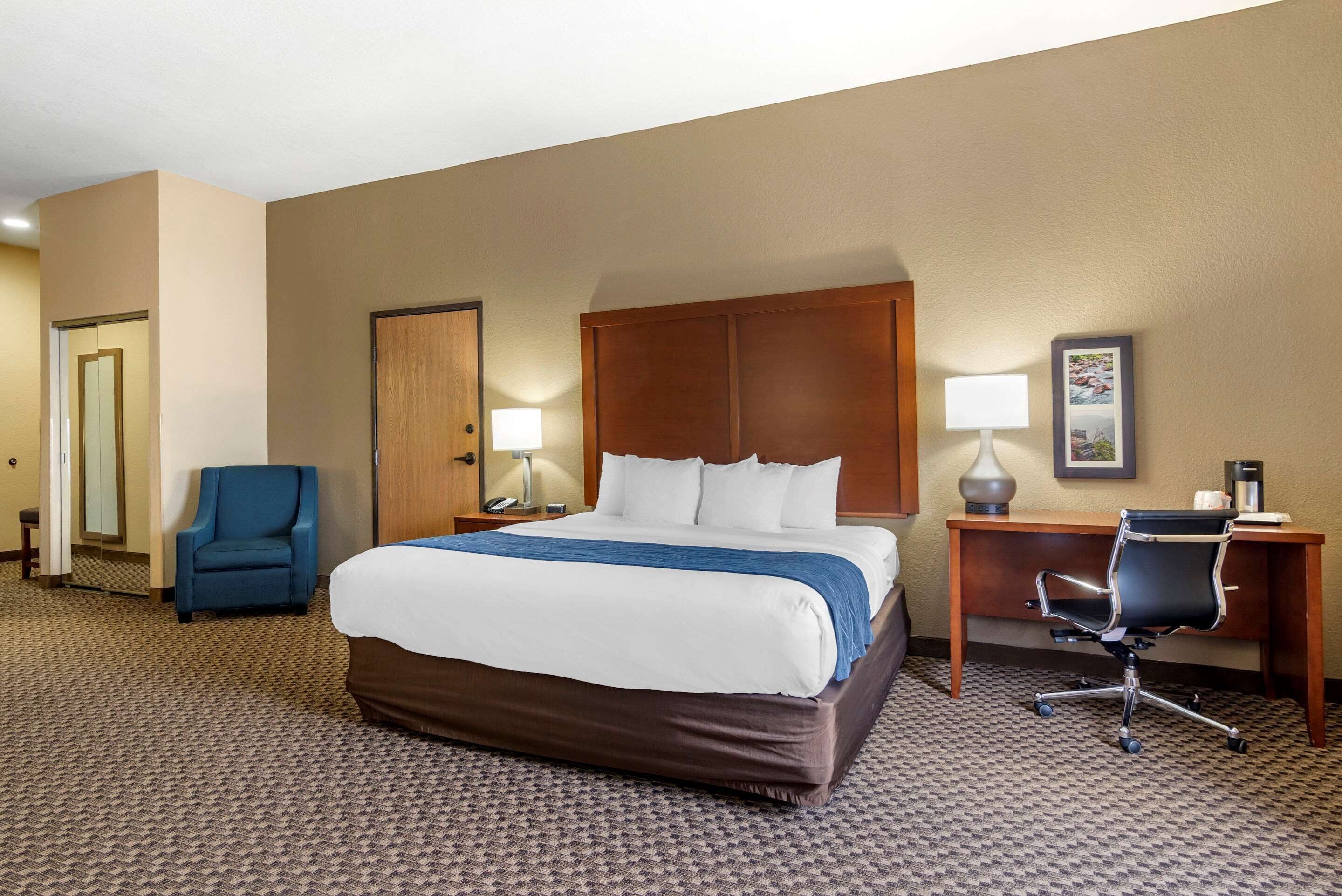 A hotel room with a large bed, a desk with a chair, a blue armchair, and a framed picture on the wall.