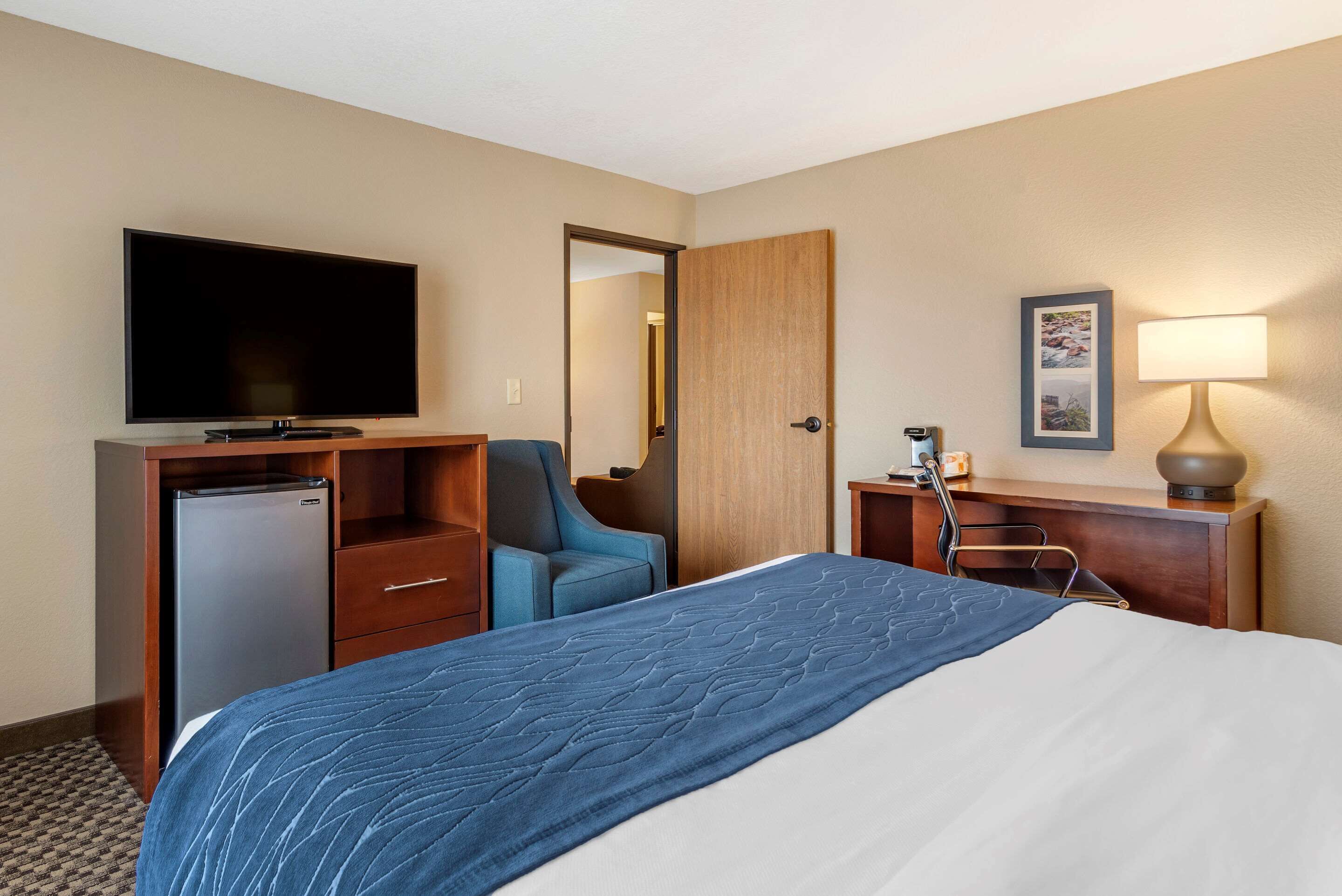 Quarto de hotel com cama, televisão, escrivaninha com abajur, cadeira e frigorífico.