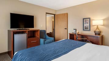 A hotel room with a bed, a television, a desk with a lamp, a chair, and a refrigerator.