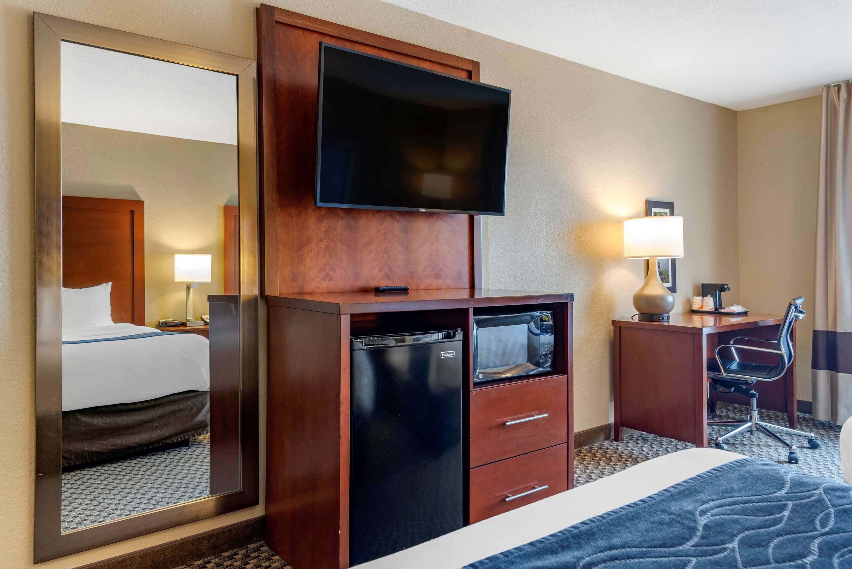 A hotel room with a bed, a TV mounted on a wooden cabinet, a microwave, a desk with a chair, and a lamp.
