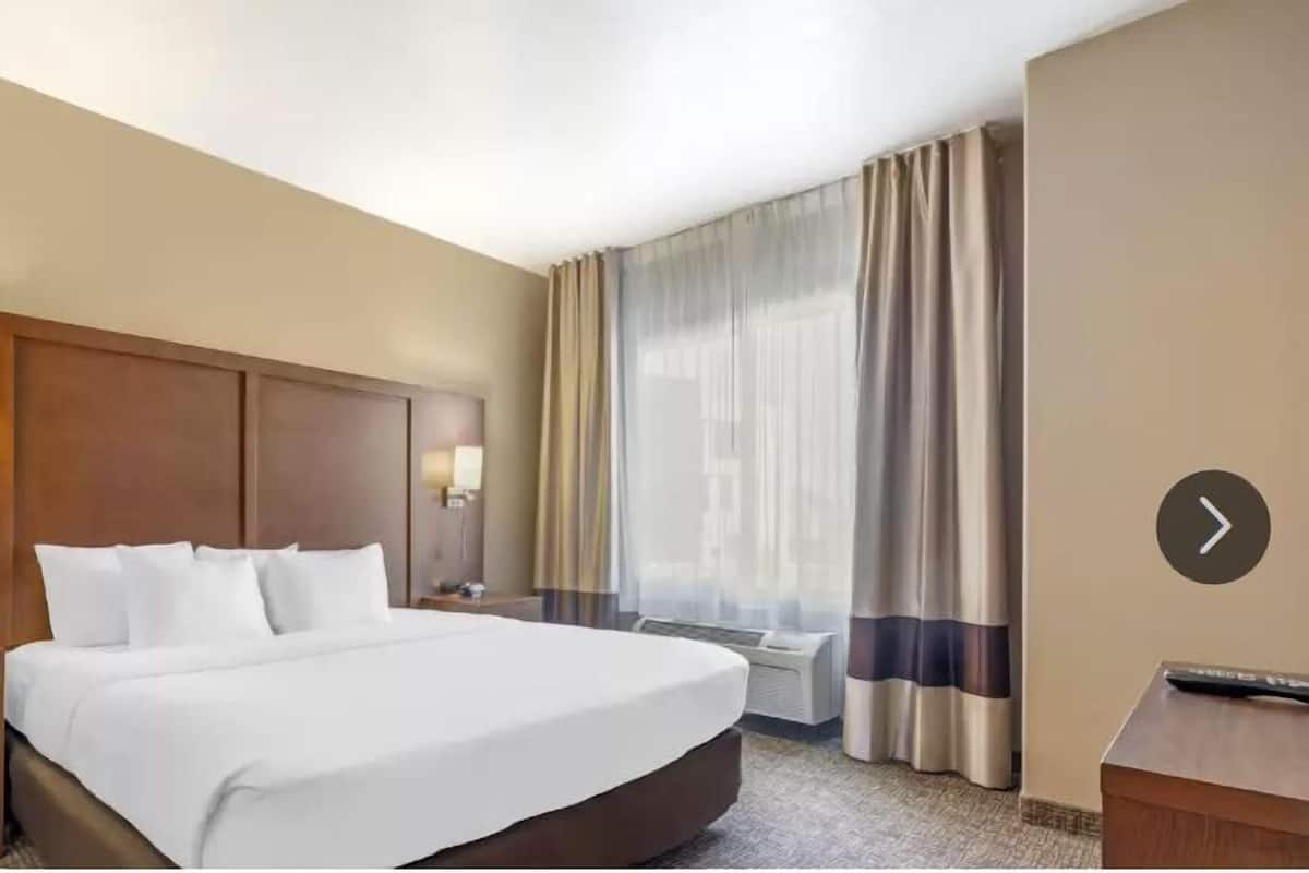 A hotel room with a large bed, a nightstand with a lamp, a desk with a remote control, and a window with curtains.